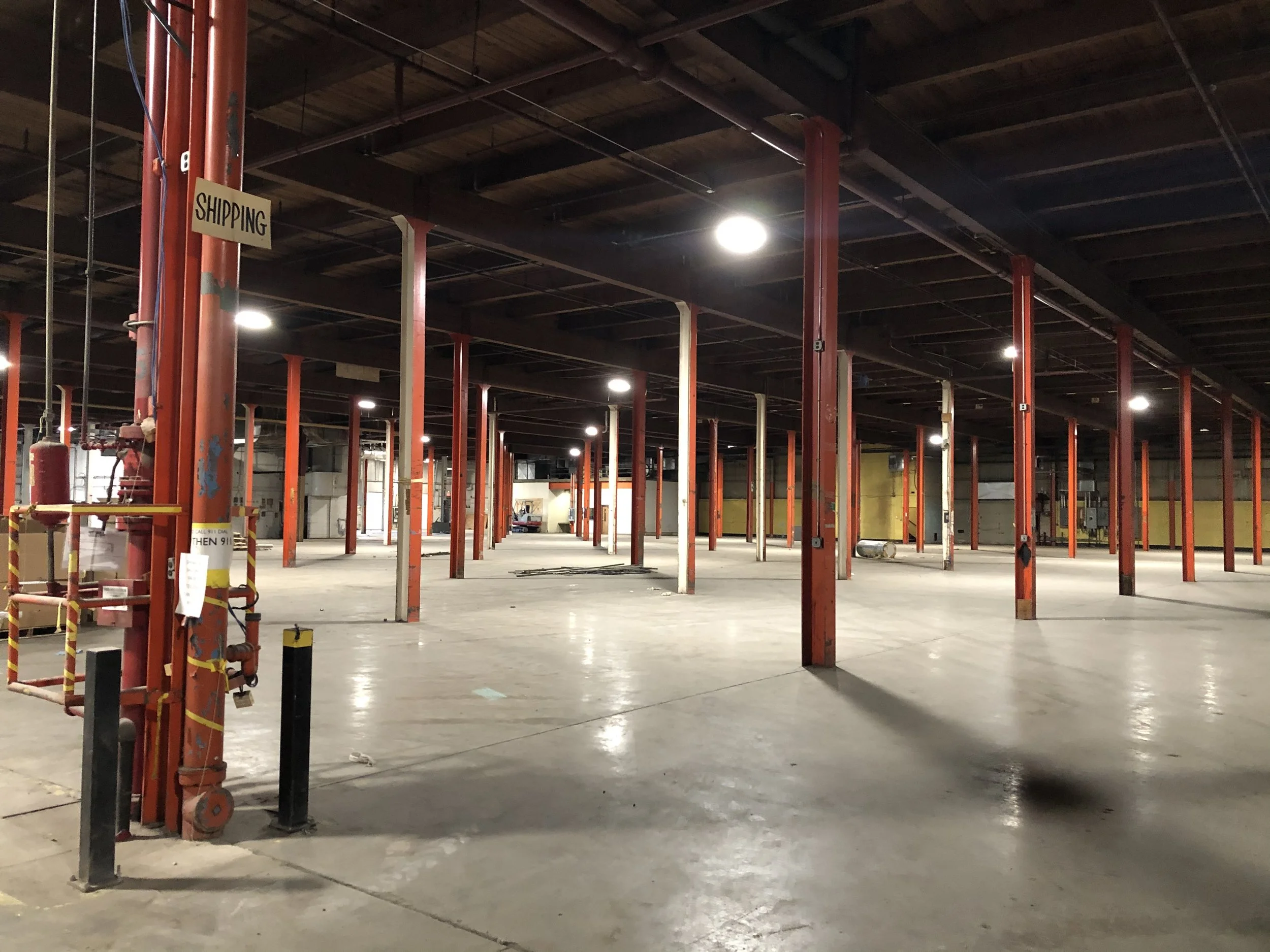 Empty warehouse or parking garage with red vertical support beams, overhead lighting, and a sign that reads 'Shipping'.