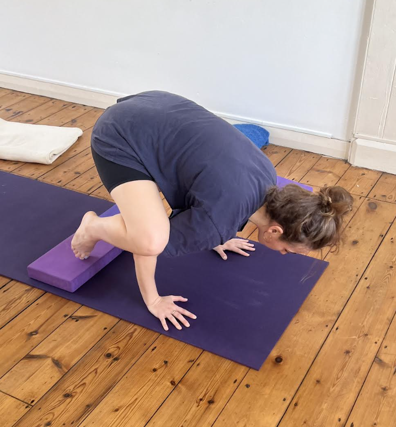 A woman practicing yoga on a purple mat, balancing with her knees on purple yoga blocks and her hands on the ground.