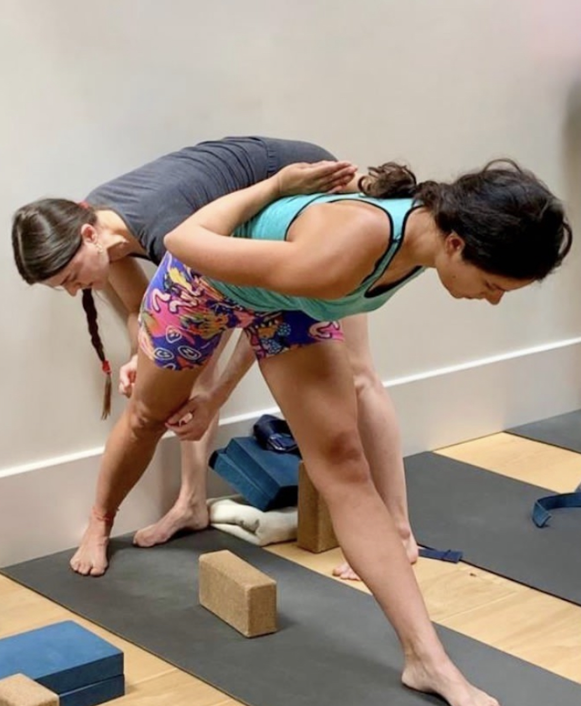 Two women practicing yoga in a studio, one in a bent-over position with one leg extended, and the other supporting her from behind.