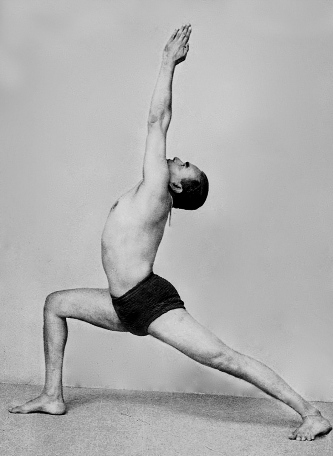 A person practicing yoga in a low lunge pose with one arm extended upward and the other arm extended downward, wearing black shorts and shirtless.