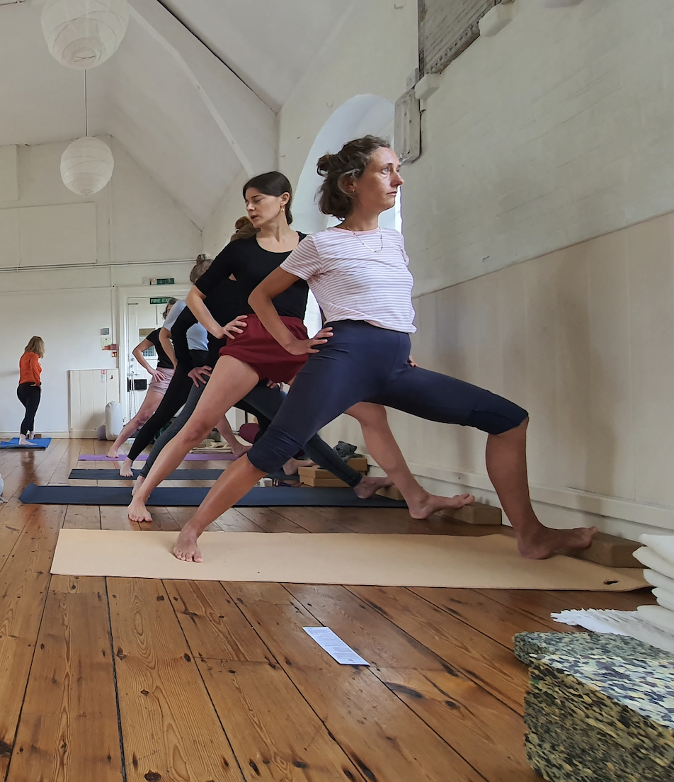 Several women practicing yoga in a studio, performing a wide-legged forward bend pose against a wall.