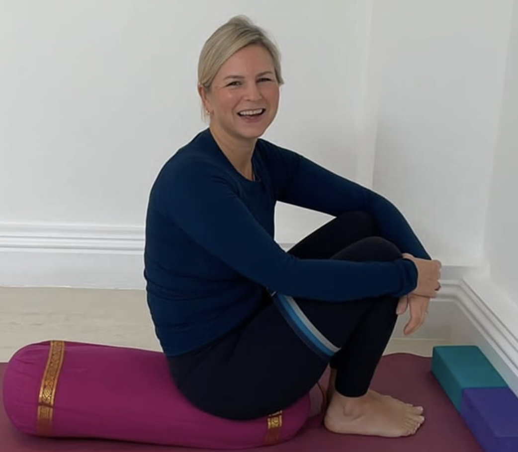 Woman sitting cross-legged on a pink yoga bolster, smiling, with yoga blocks nearby, indoors against a white wall.