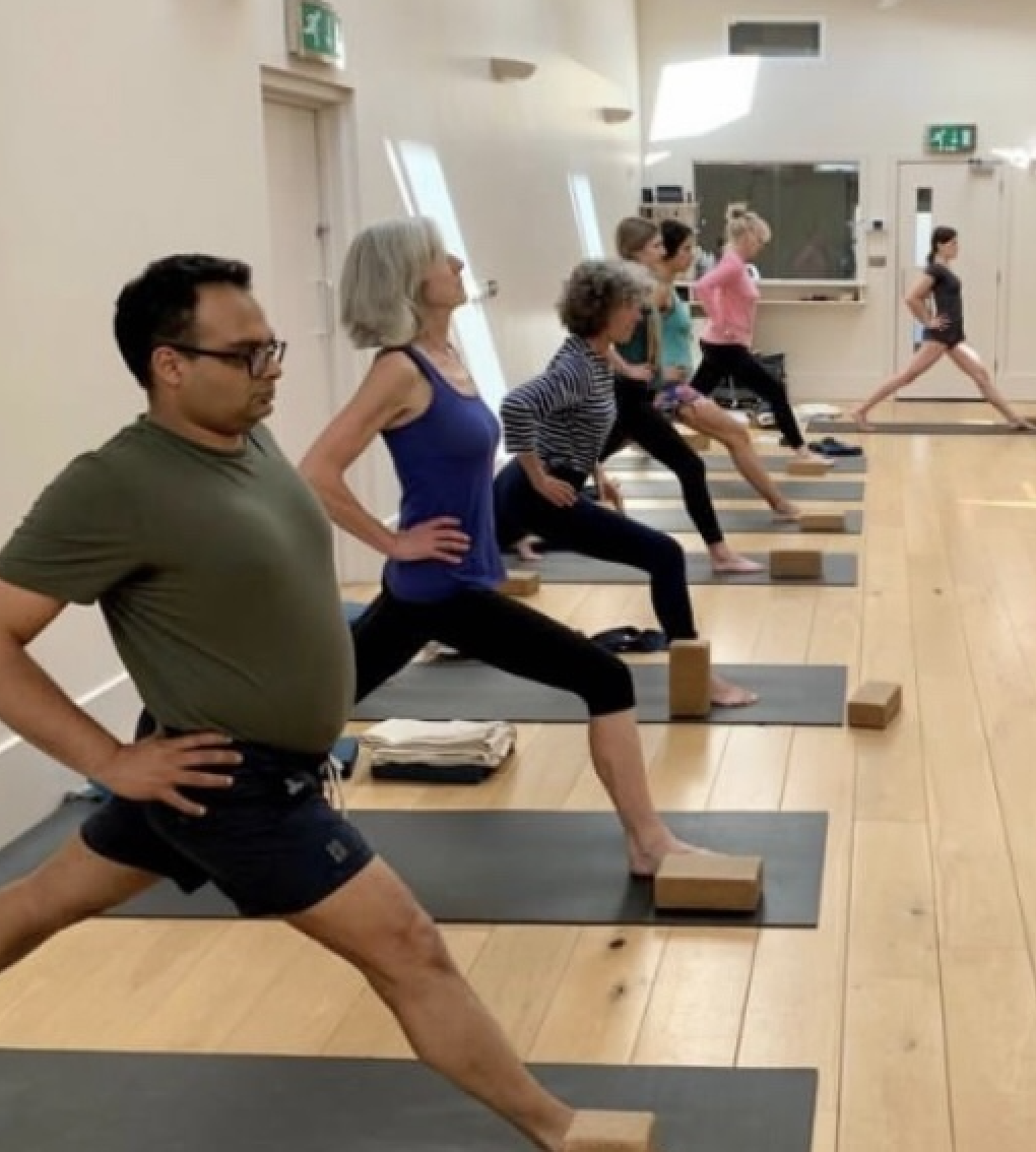 A group of people in a yoga class, performing lunges with yoga blocks under their front feet in a spacious, well-lit room.