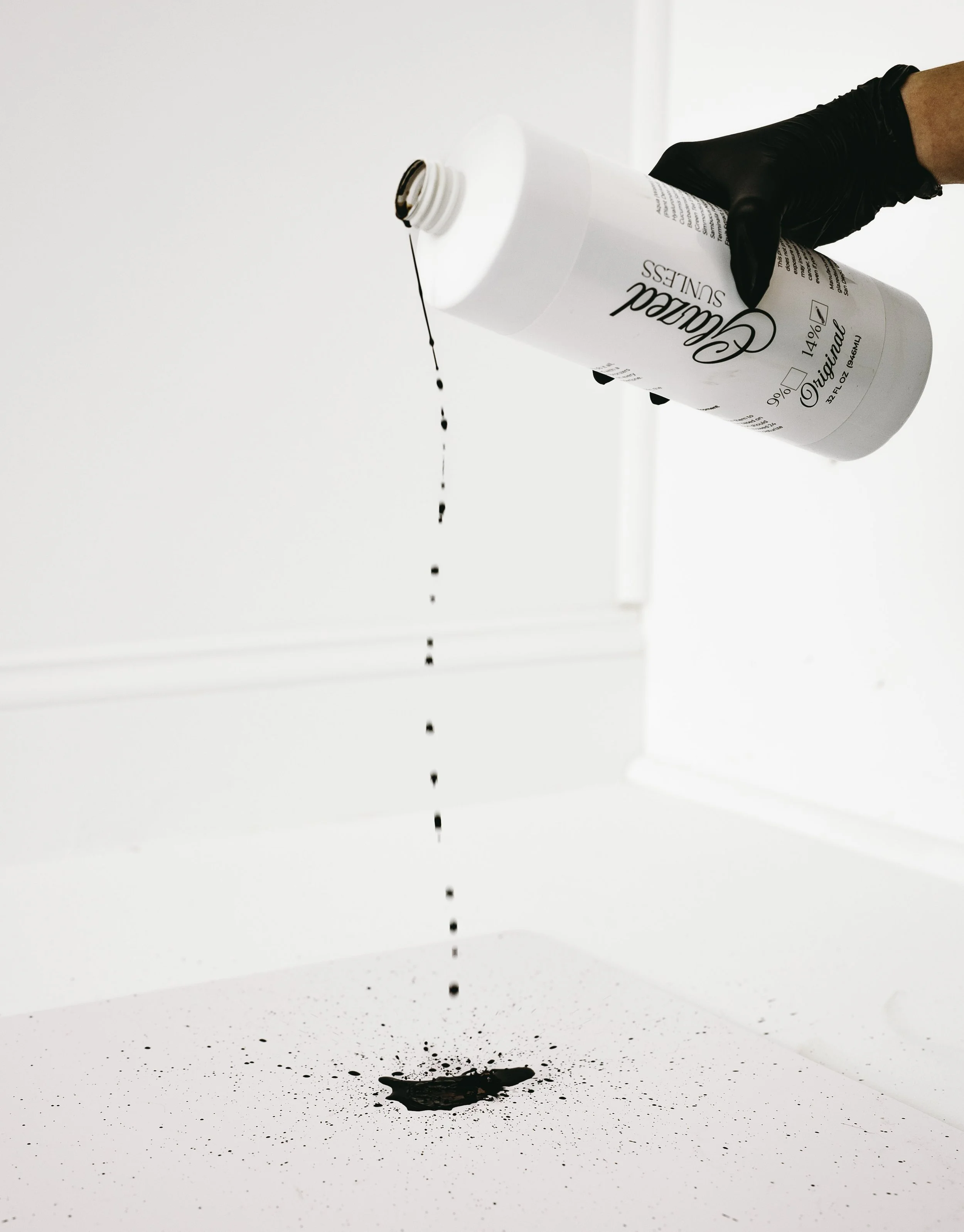 A hand wearing a black glove is pouring black ink from a white bottle onto a white surface. The ink is dripping down and splattering on the surface below.