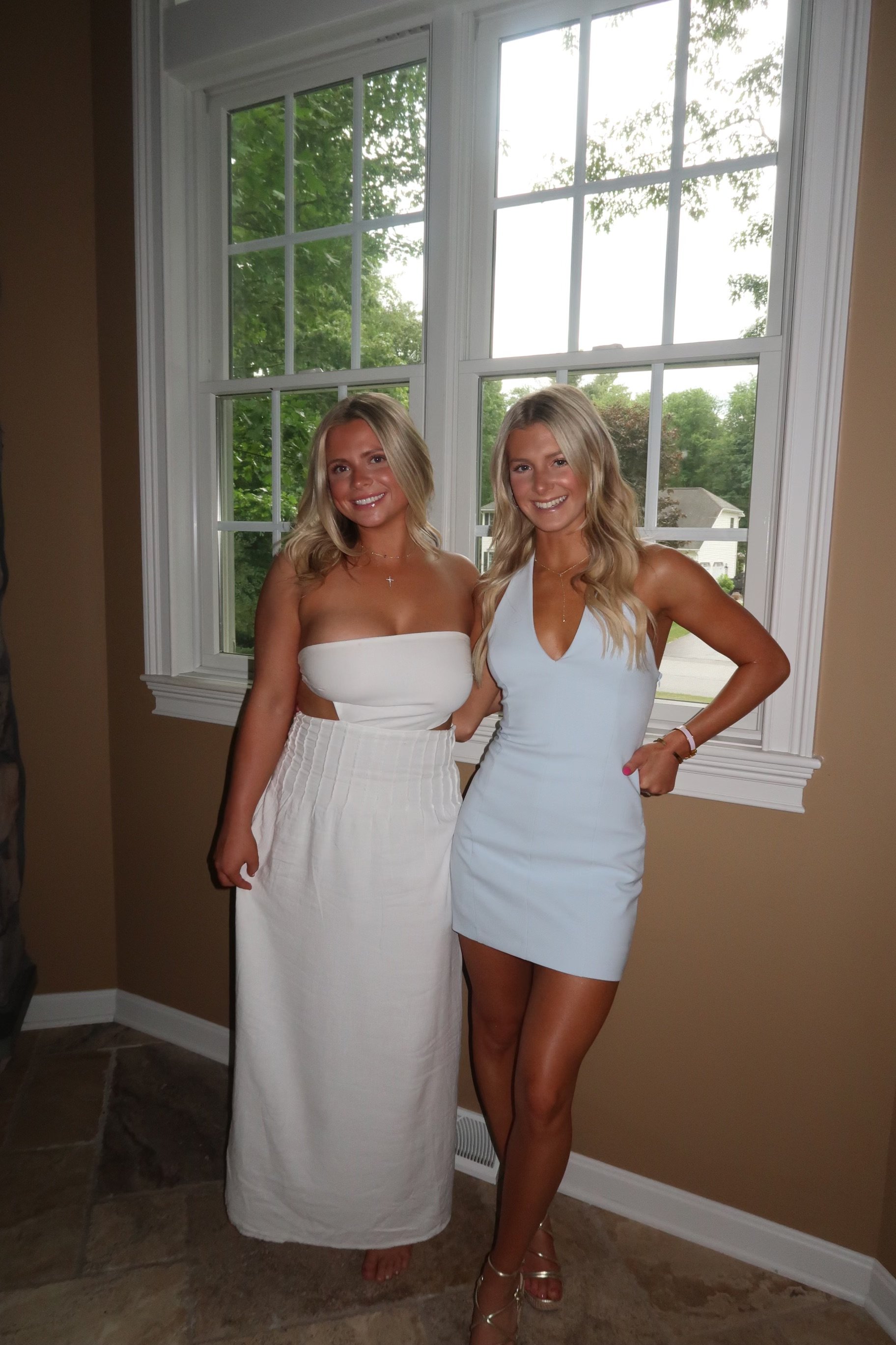 Two young women with blonde hair and light skin standing next to each other in front of a large window, smiling and dressed in white summer dresses, with one wearing silver heels and the other barefoot.