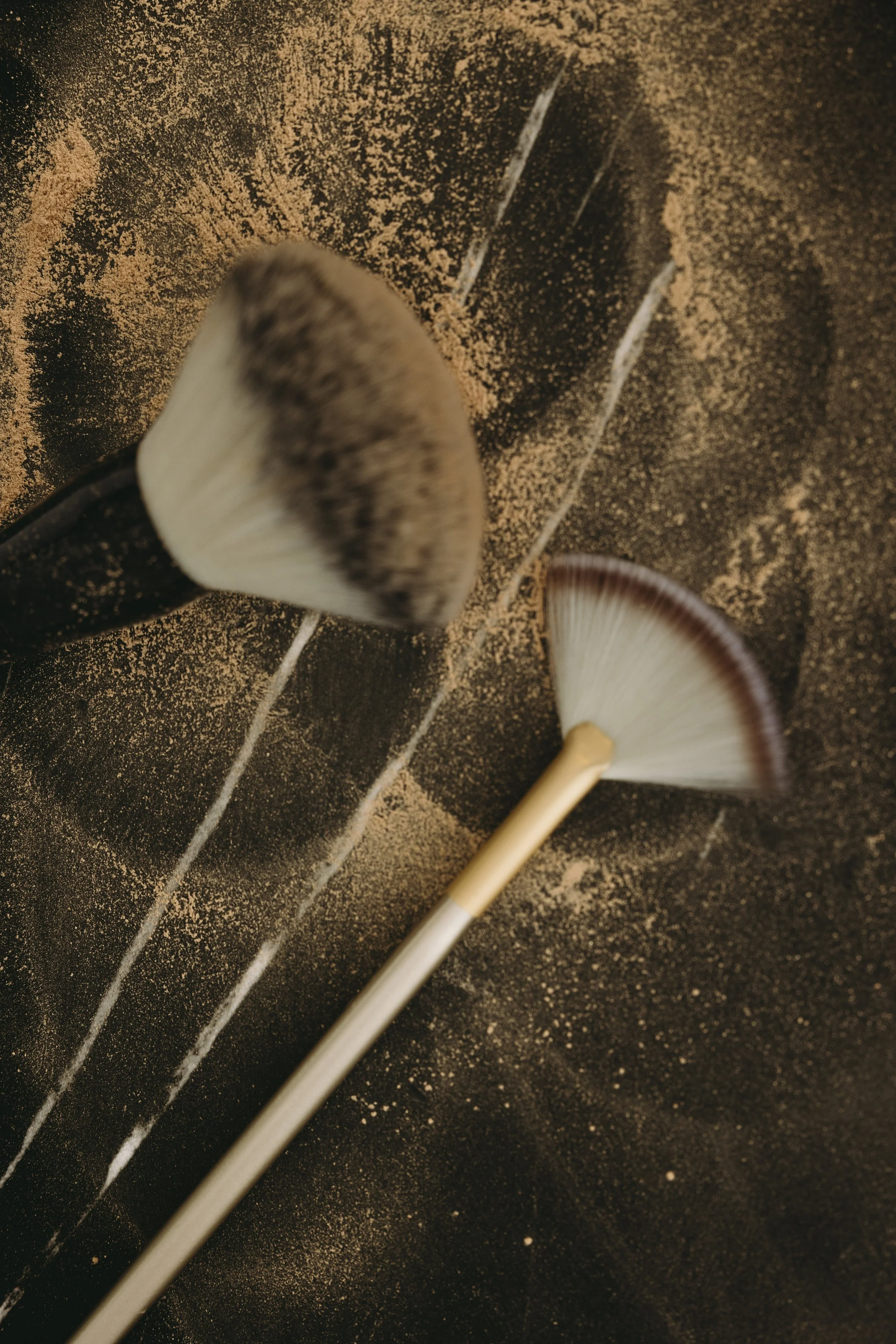 A makeup brush dusted with a light brown powder, with more brown powder scattered on a dark surface. Another makeup brush is partially visible in the bottom right.