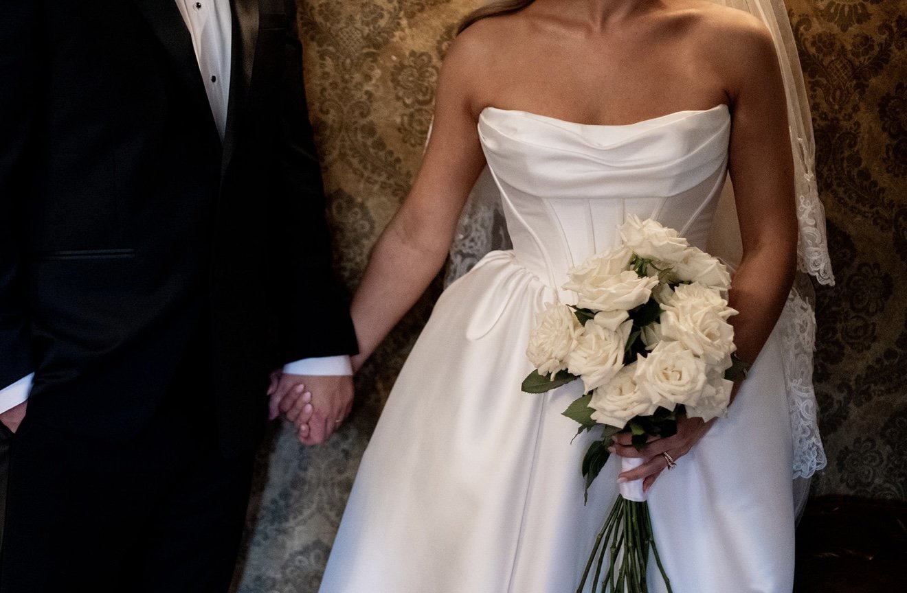 A bride and groom holding hands on their wedding day. The bride is wearing a strapless white wedding gown and holding a bouquet of white roses, while the groom is dressed in a black tuxedo with a white shirt.