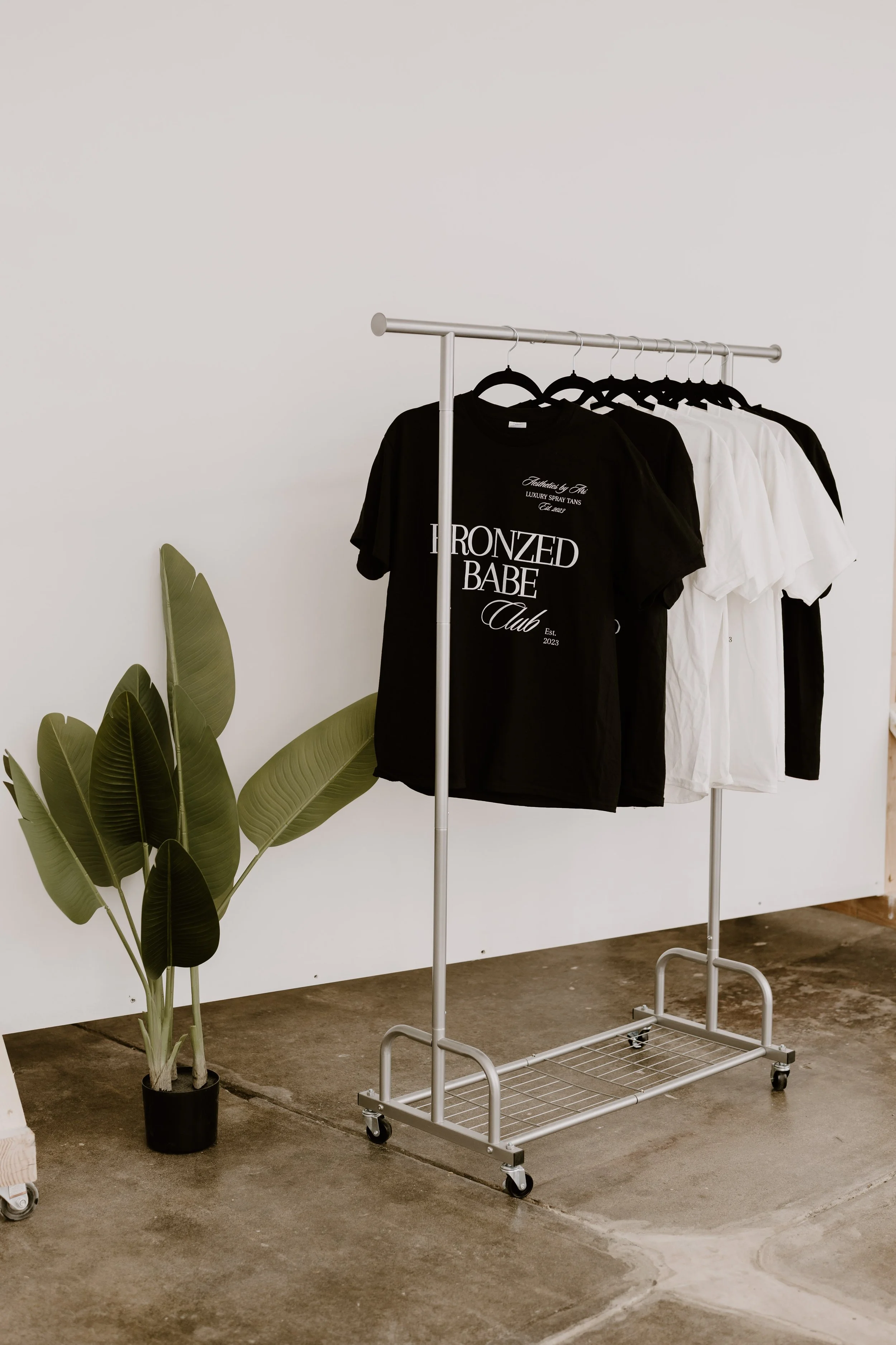 A clothing rack on wheels holding black and white T-shirts with printed text. To the left, there is a potted plant with large green leaves. The scene appears to be in a minimalist indoor space with a concrete floor and a white wall.