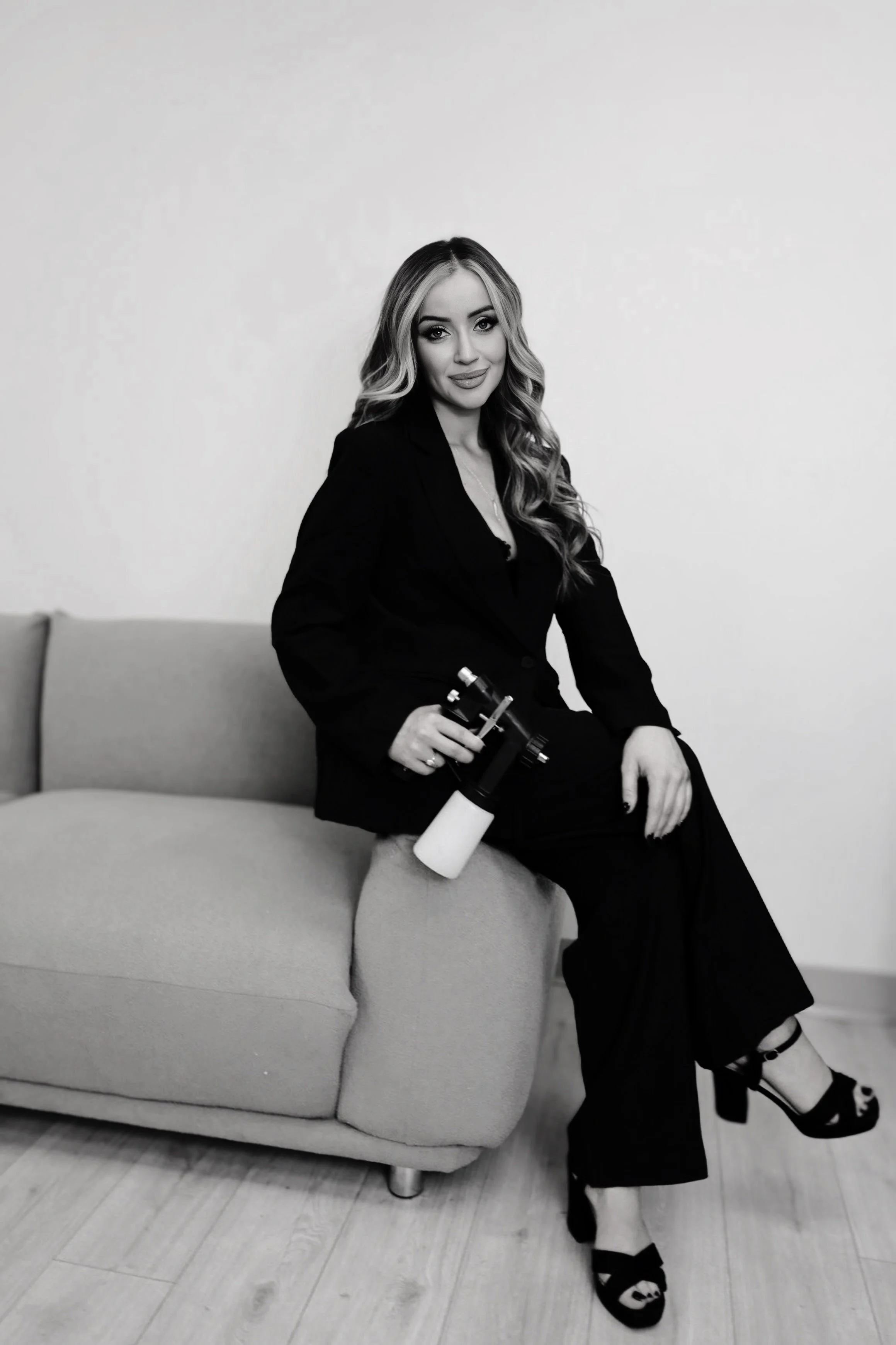 A woman with long wavy hair, wearing a blazer and dark pants, sitting on a sofa and holding a telescope.