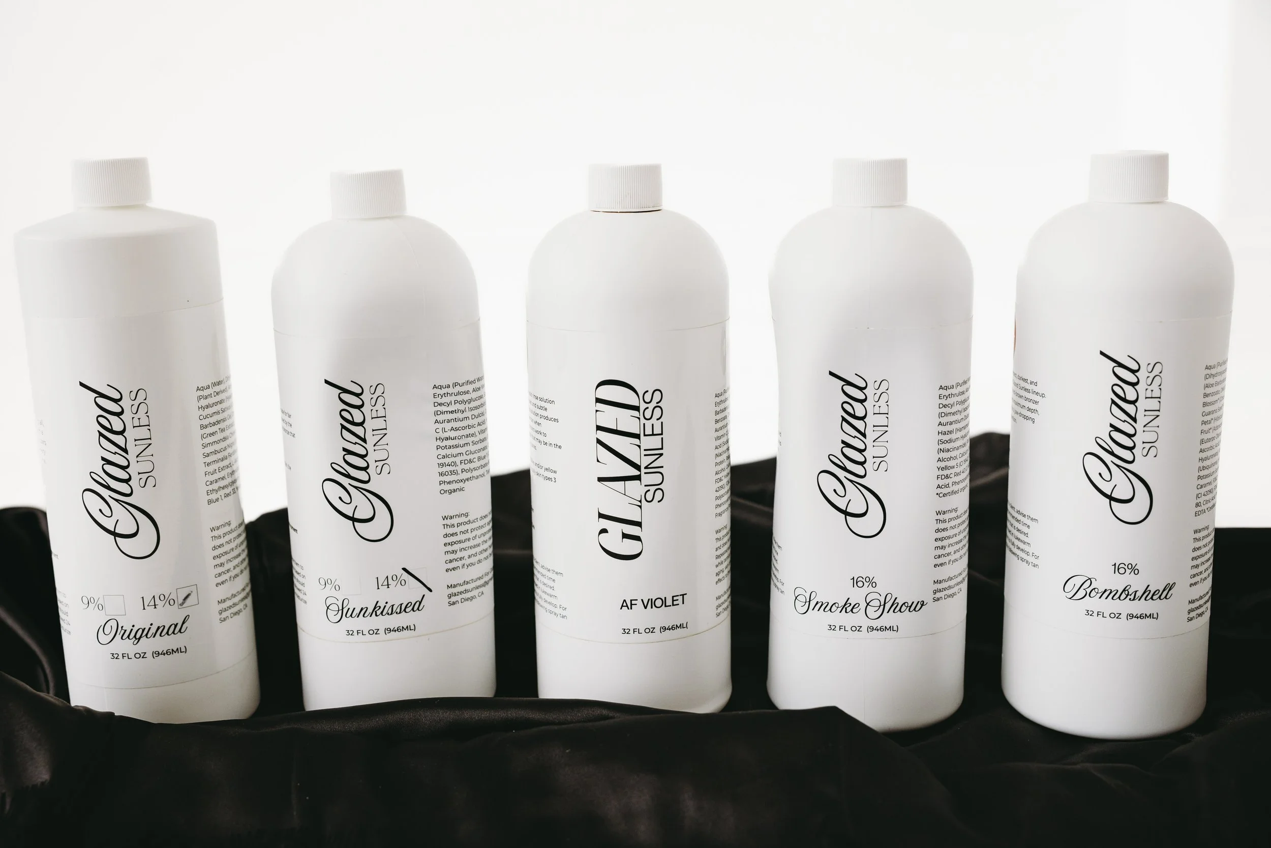 A lineup of five white bottles of sunscreen with black cursive text, displaying different SPF percentages, placed in a black container against a plain light background.