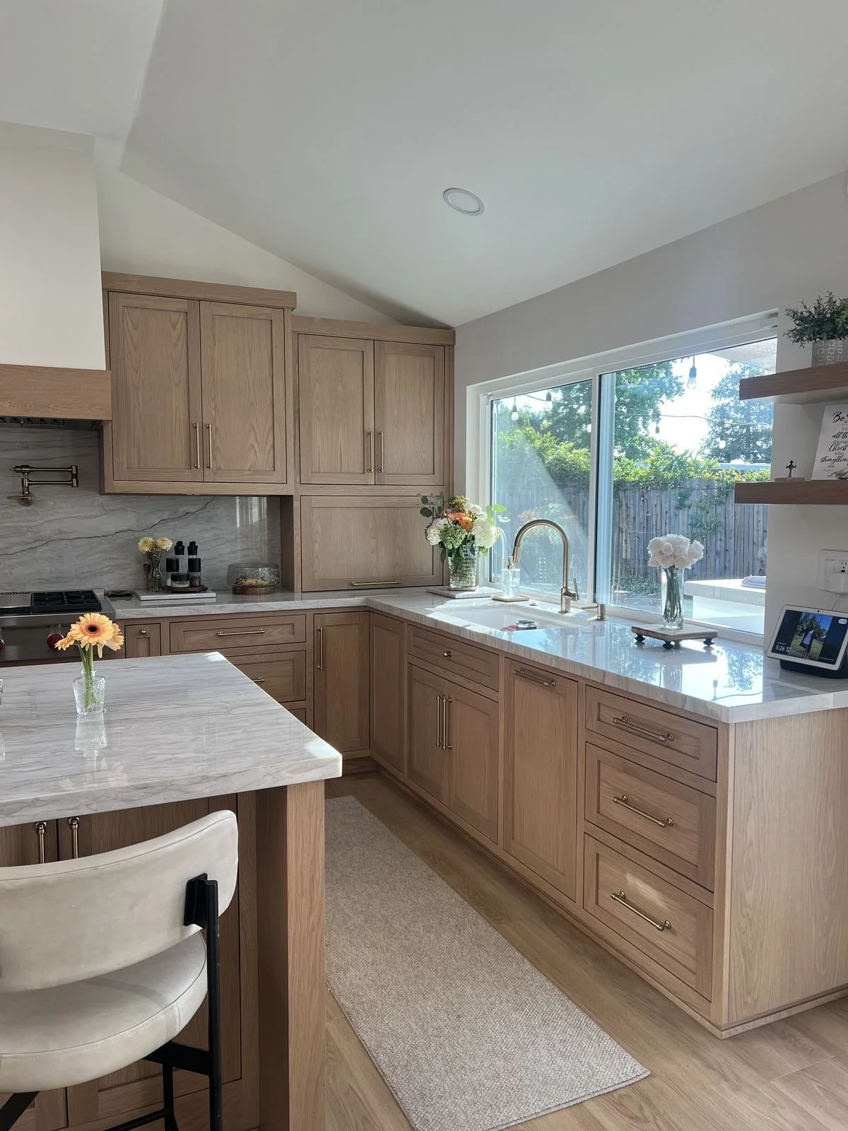 A bright kitchen with light wood cabinets, white marble countertops, a large window with a view of the backyard, and small vases with flowers on the countertops and island.