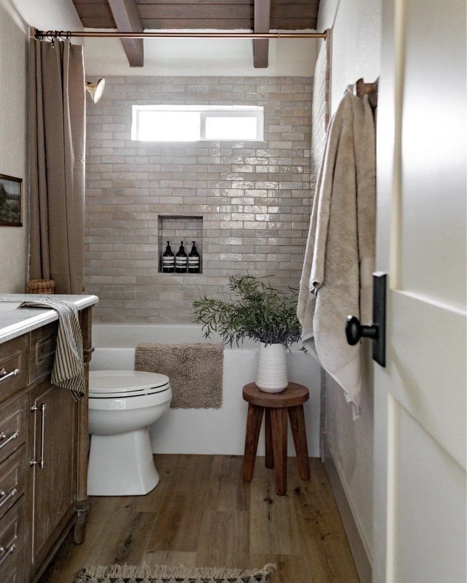 A small modern bathroom featuring a white toilet, a wooden vanity with a countertop, beige towels, a potted plant on a small wooden stool, and a brick shower wall with built-in shelves holding black bottles. The bathroom has wooden ceiling beams, a narrow horizontal window, and a wooden floor.