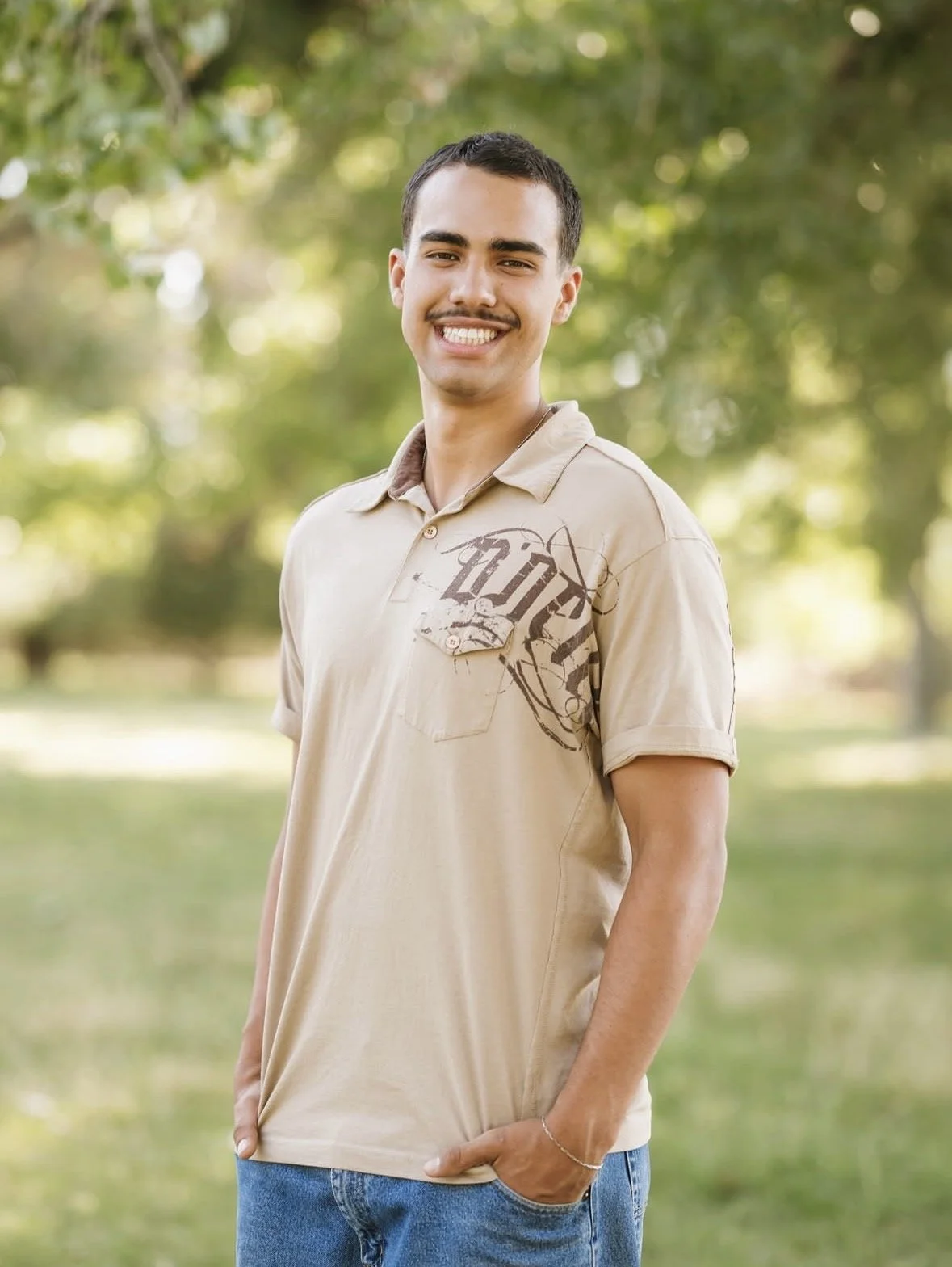 A young man smiling outdoors in a park. He is wearing a beige polo shirt and blue jeans, with his right hand in his pocket.