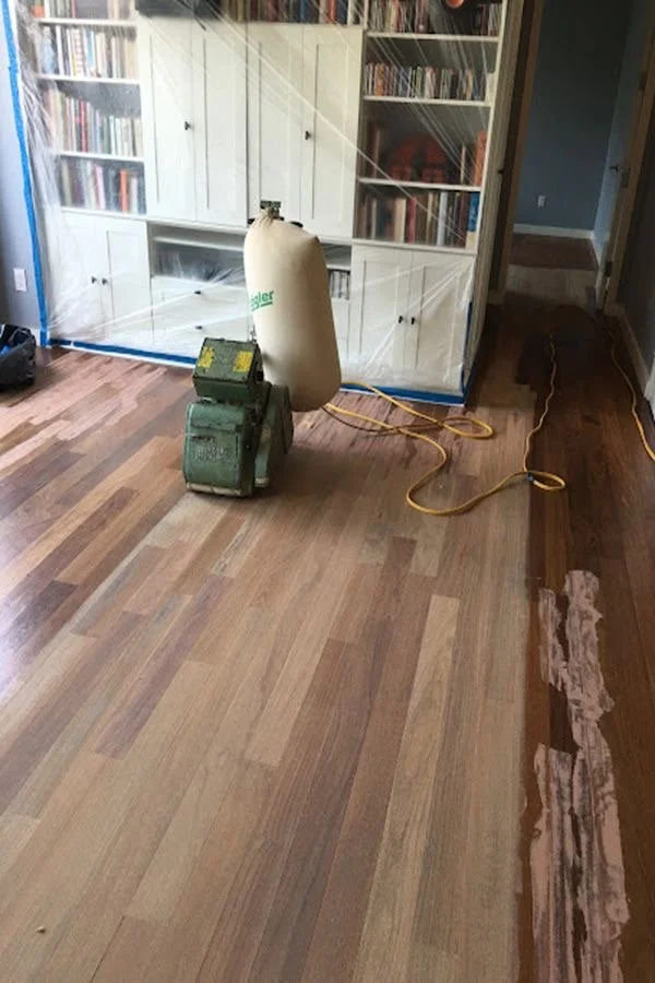 A floor sander and a large white dust bag setup in a room with a wooden floor, next to a white bookcase and a blue wall, with a cord running across the floor.