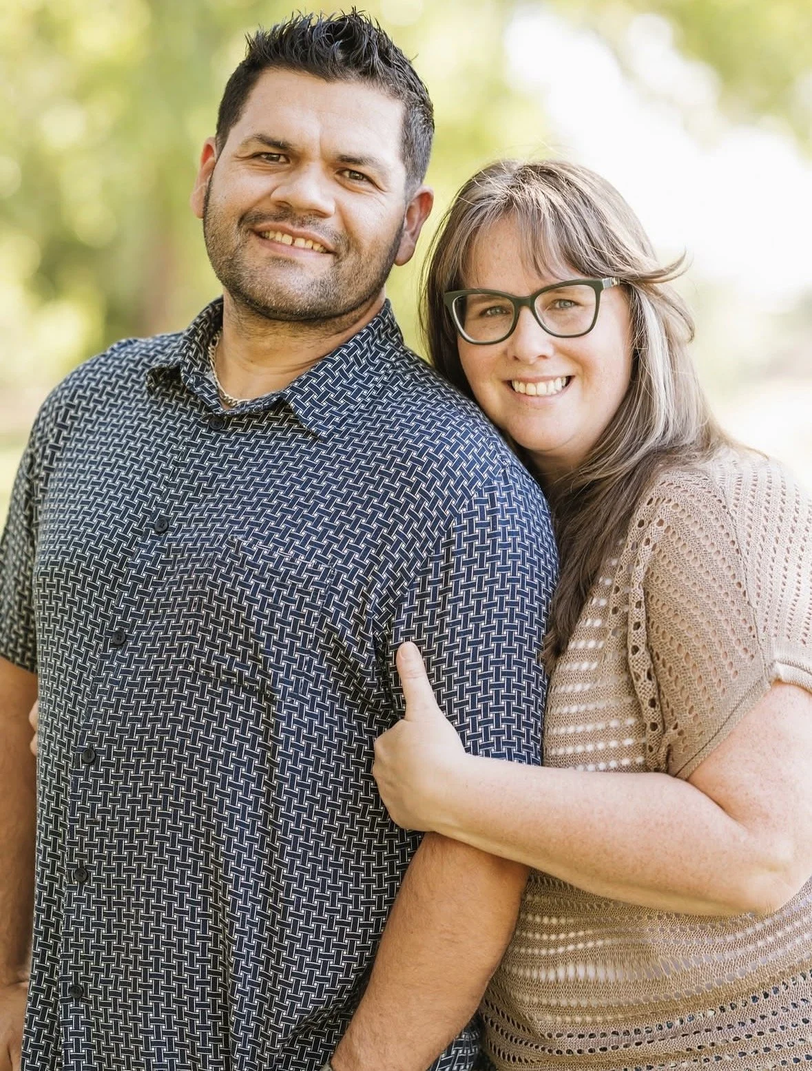 A man and a woman standing outdoors, smiling, with the woman embracing the man's shoulder. The man has dark hair, a beard, and is wearing a patterned button-up shirt. The woman has long hair, glasses, and is wearing a light-colored, short-sleeved sweater.