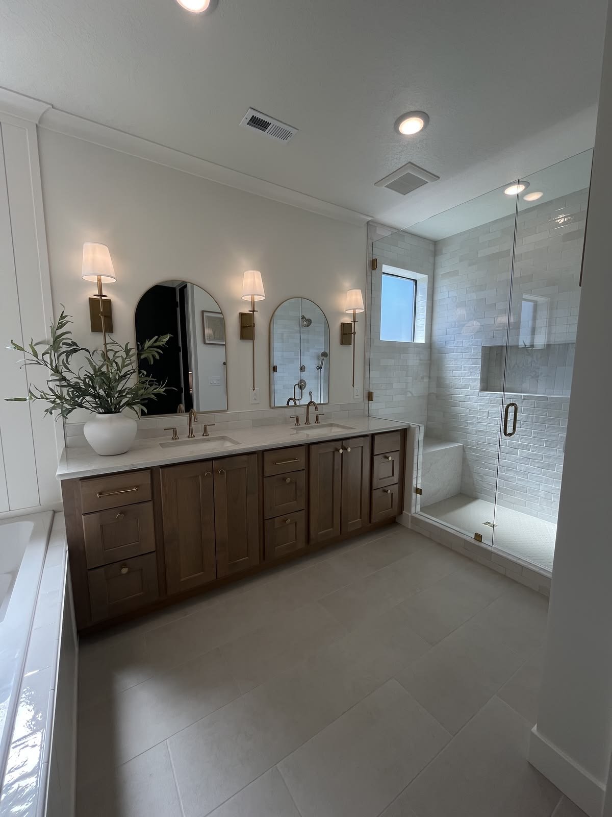 Modern bathroom with dual sinks, wooden vanity, wall-mounted lights, a glass-enclosed shower with white brick tile, a small window, and a white vessel sink with green plant decor.