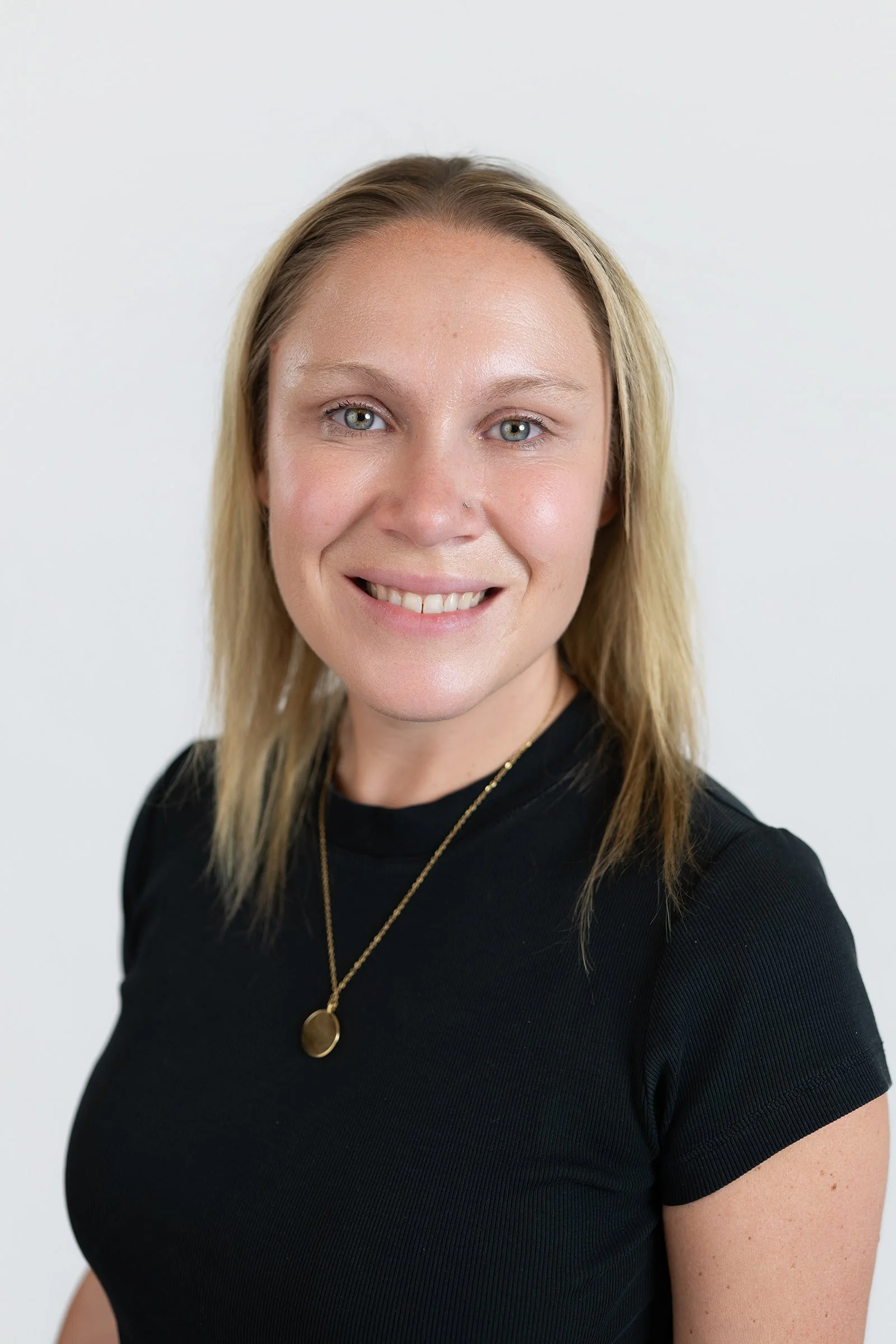 A woman with blonde hair and light skin smiling, wearing a black top and gold jewelry, standing against a plain white background.