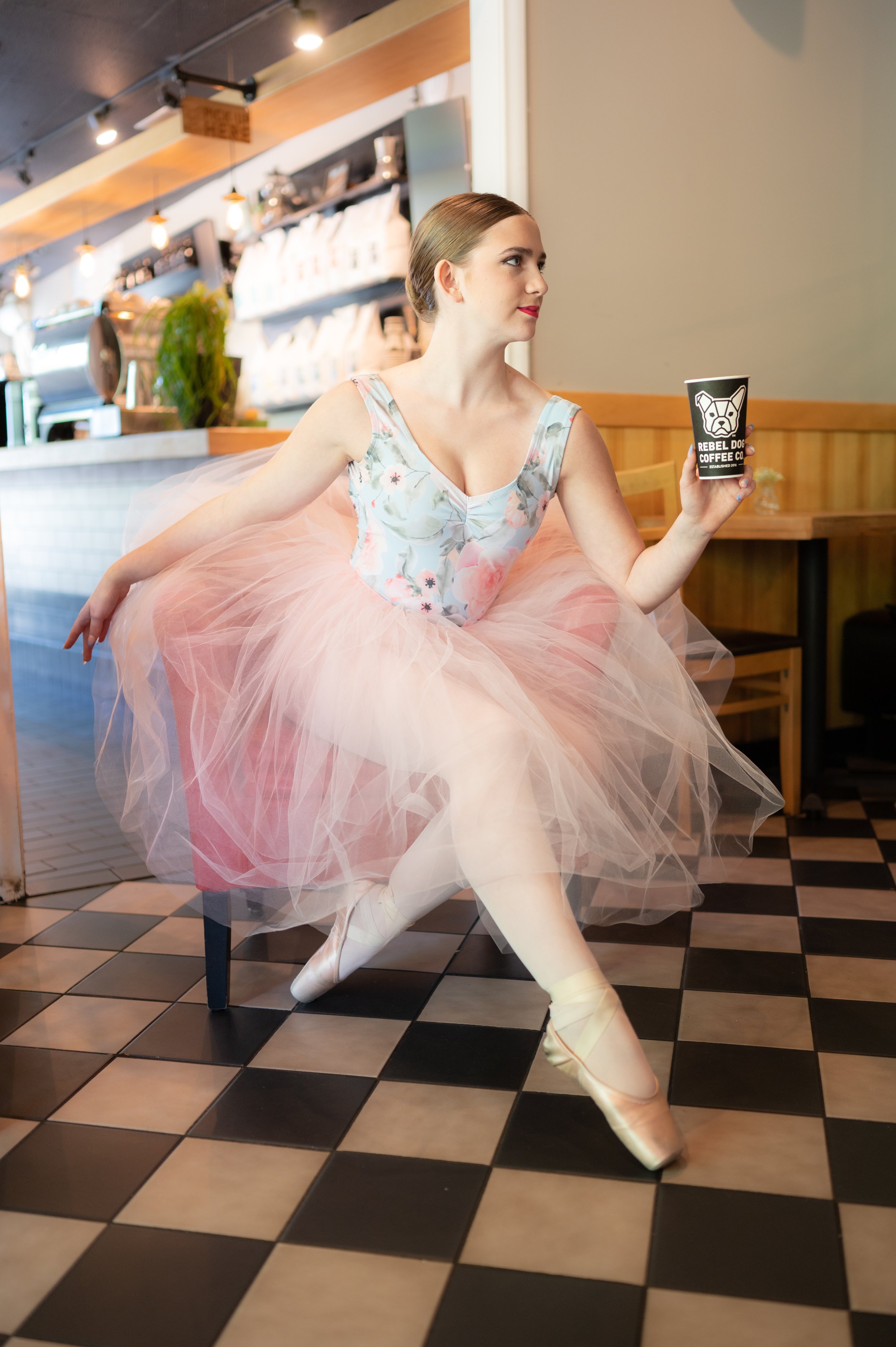 A ballet dancer sitting on a chair in a coffee shop, holding a cup with a dog logo. She is wearing a floral ballet costume with a pink tutu, has her hair in a bun, and is wearing ballet slippers.