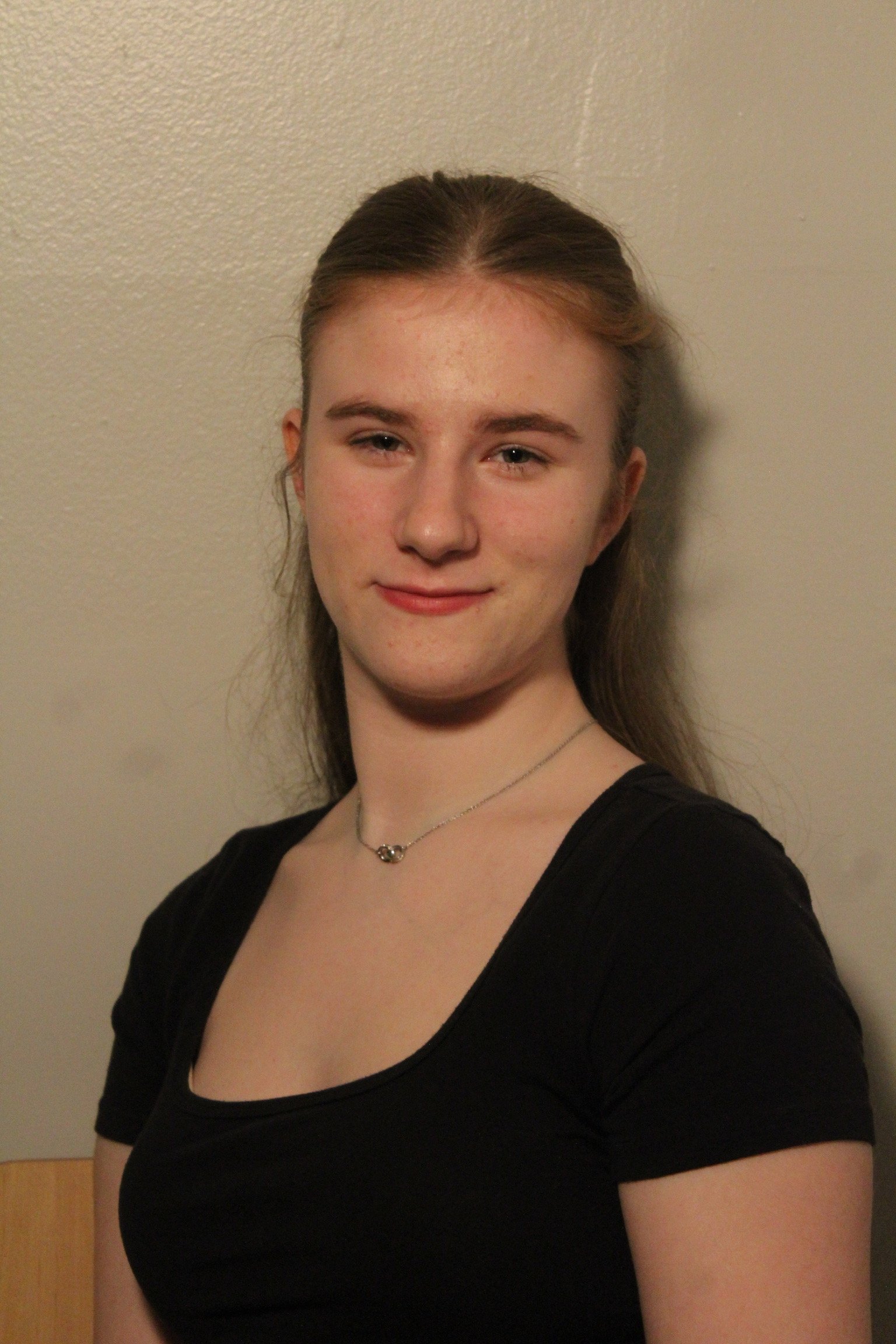 Young woman with long brown hair wearing a black top and a silver necklace, standing against a beige wall.