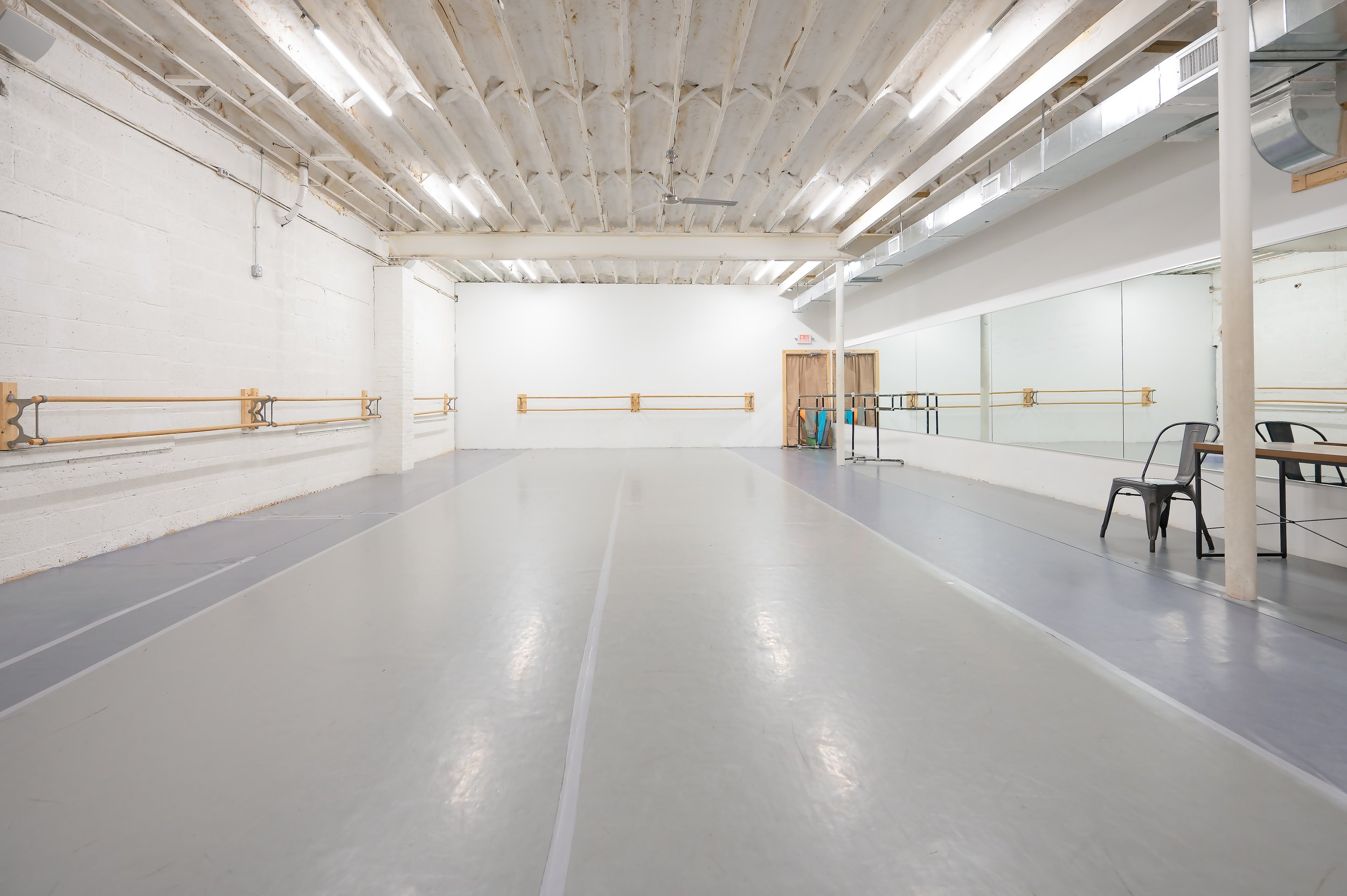 Empty dance or ballet studio with wooden ballet barres, large mirror, and gray flooring in a spacious, white-walled room with an industrial ceiling.