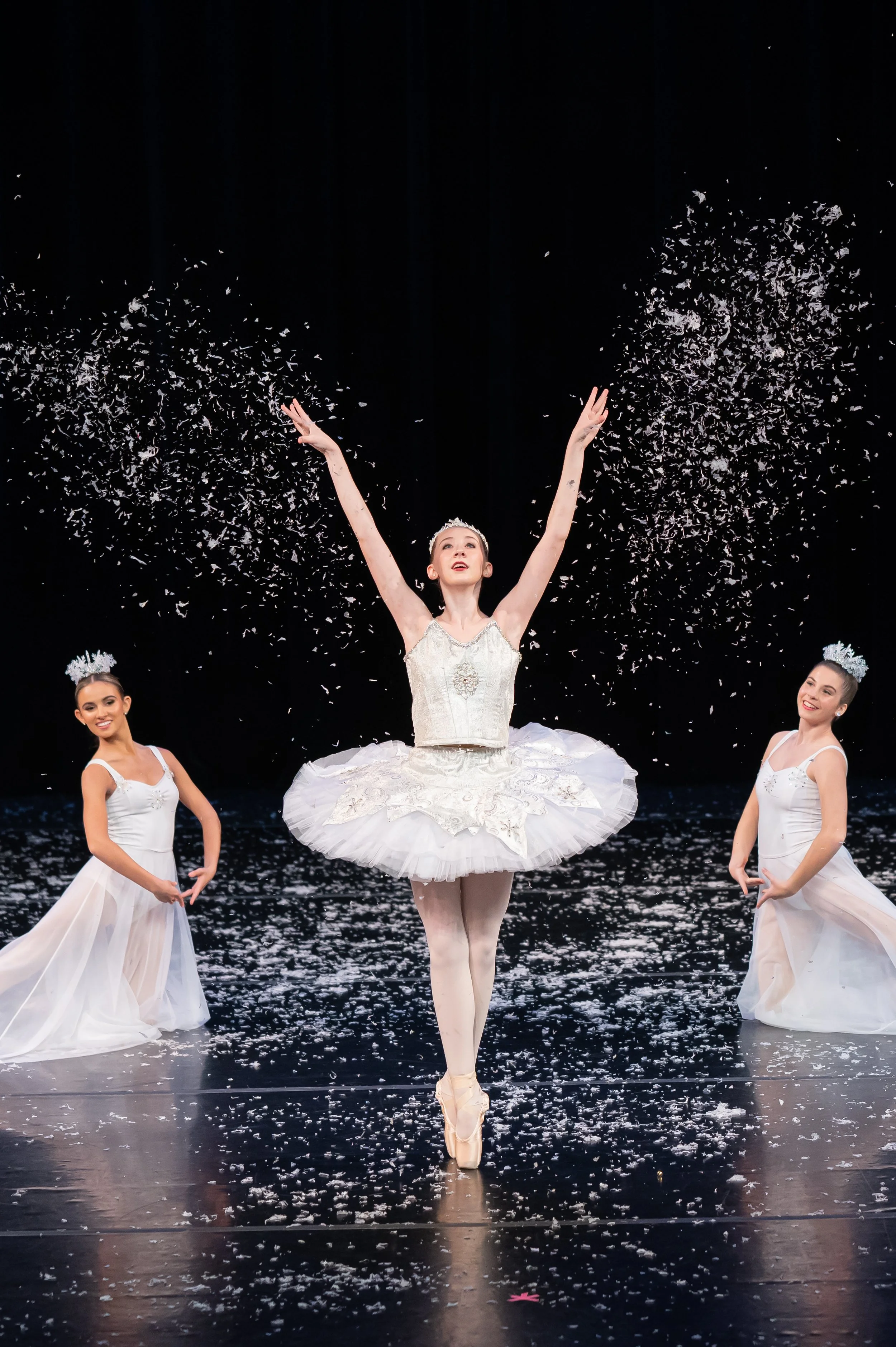 Ballet dancers on stage, with one ballerina in the center performing en pointe with arms raised, wearing a white tutu. Two other dancers in white costumes on either side, and confetti falling from above.