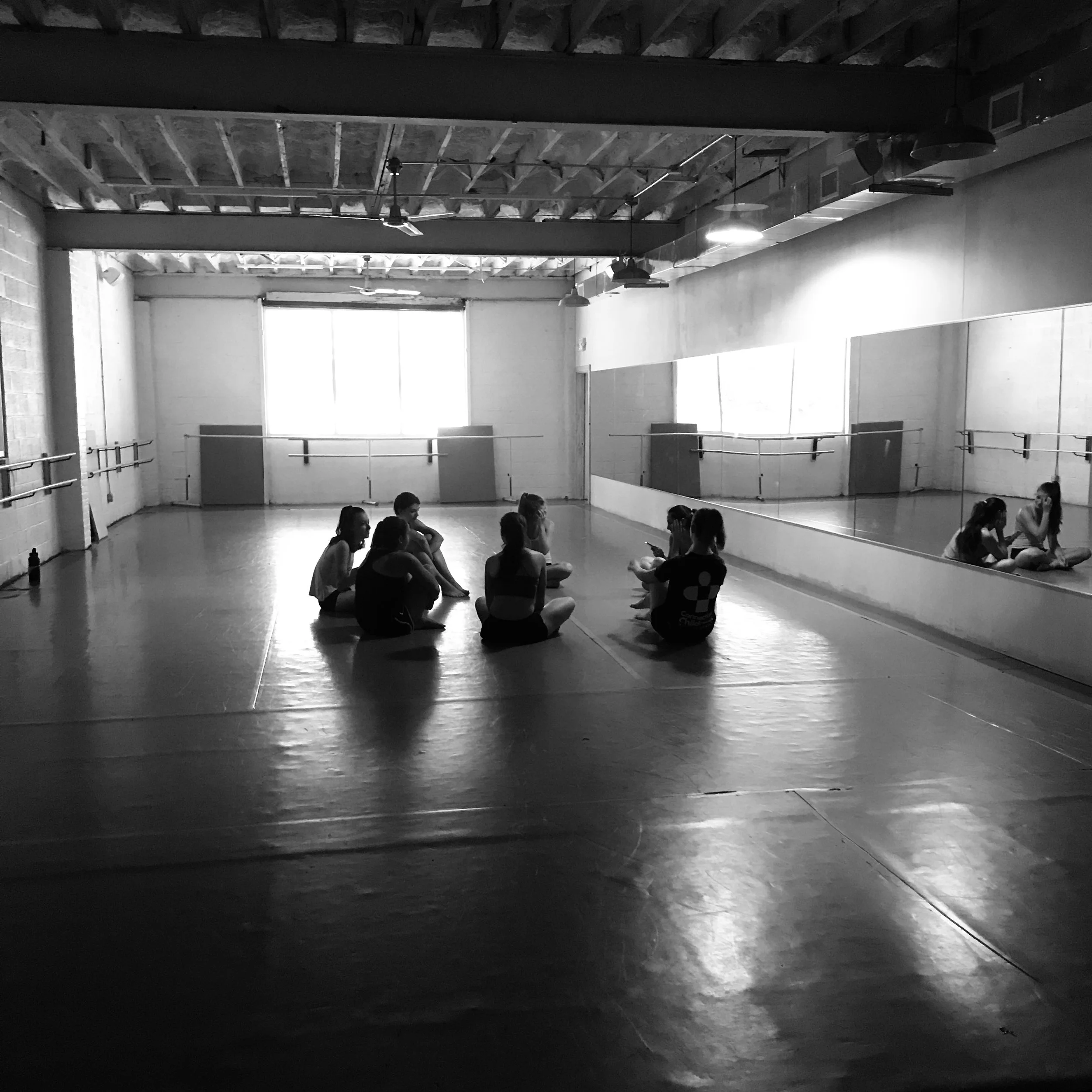 Dance studio with large mirror on the wall, ballet barre, and hardwood floor, children sitting on the floor in a row, some looking at their phones.