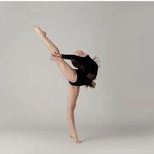 A woman in a black leotard performing a handstand with her legs extended backward in a gym or studio with a plain gray background.