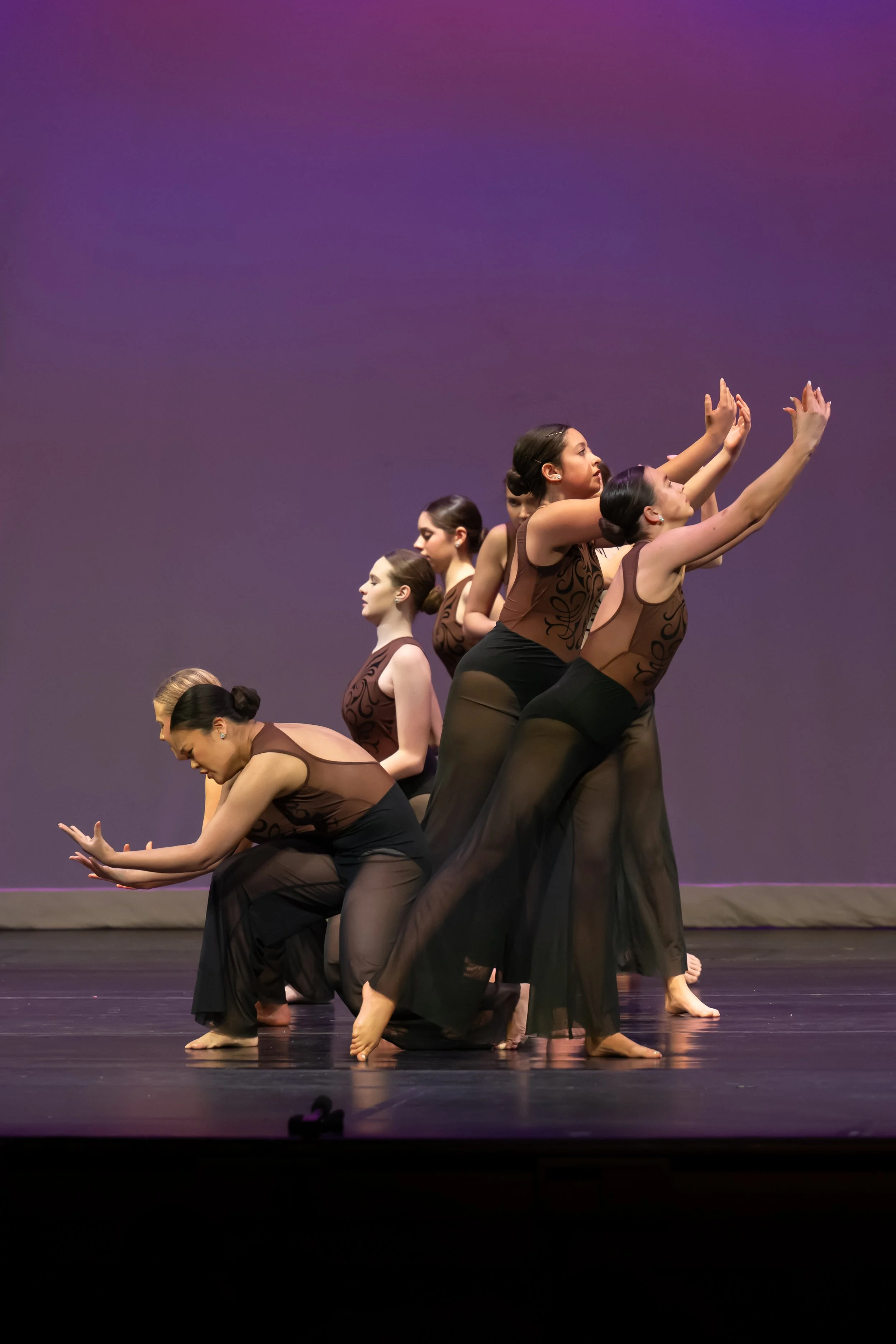 A group of female dancers wearing black sheer skirts and brown tops are performing on stage, all in a synchronized pose with their arms extended forward and heads slightly tilted upward.