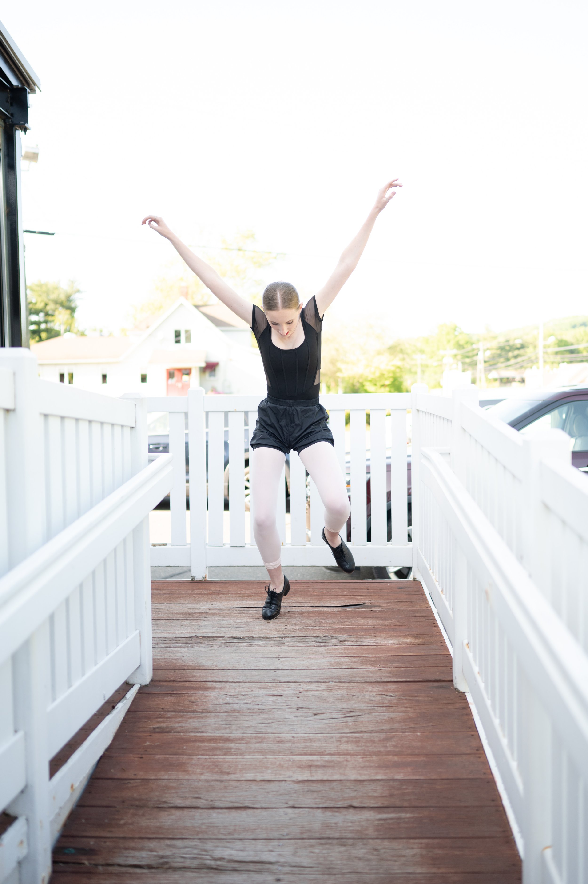 A ballerina in a black leotard and white tights practicing dance moves outdoors on a wooden deck with white fences, with houses and trees visible in the background.