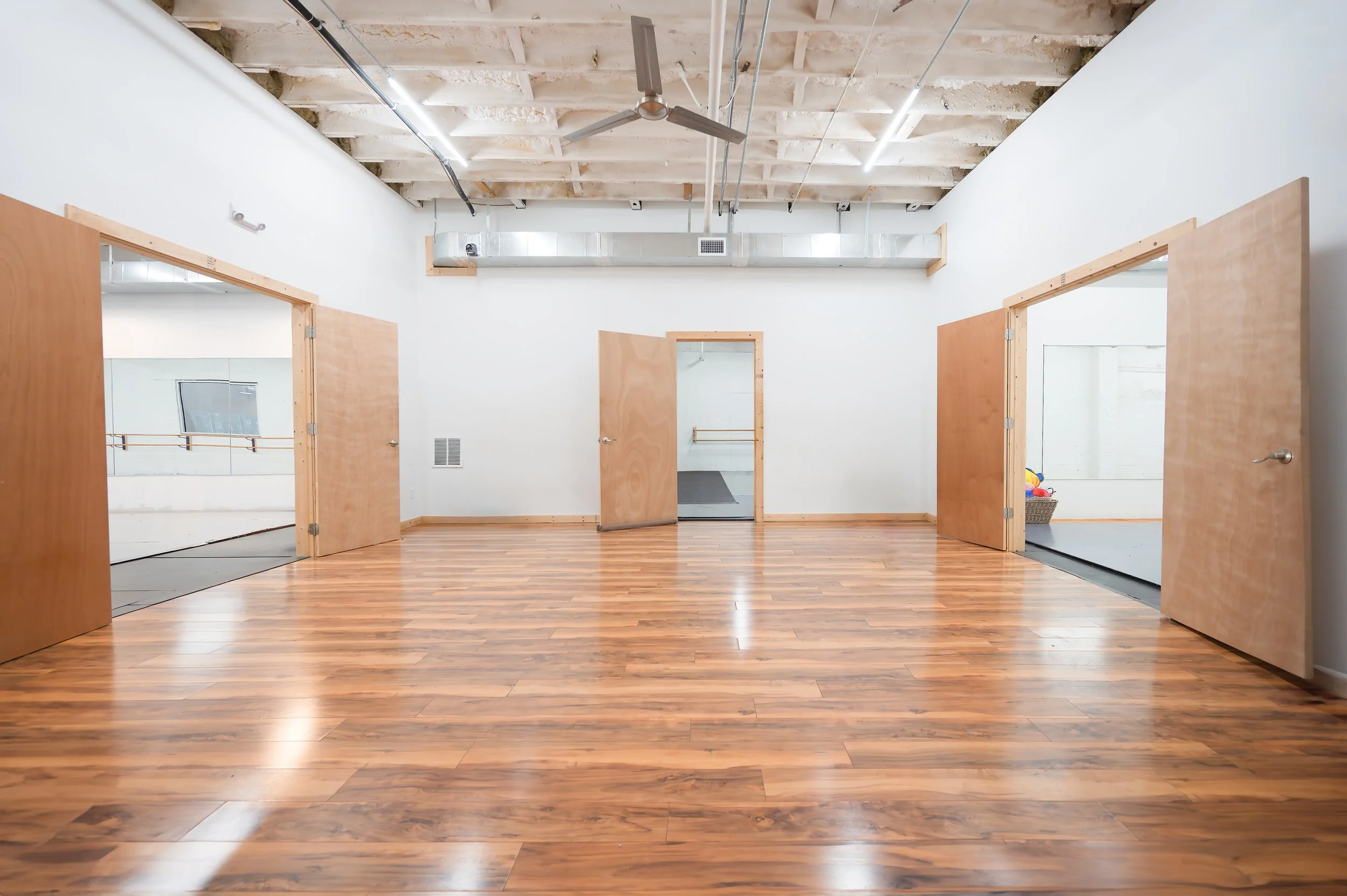 A dance studio with wooden floors, white walls, and open doorways that lead to other rooms. The ceiling has exposed beams and ductwork, with a ceiling fan in the center.