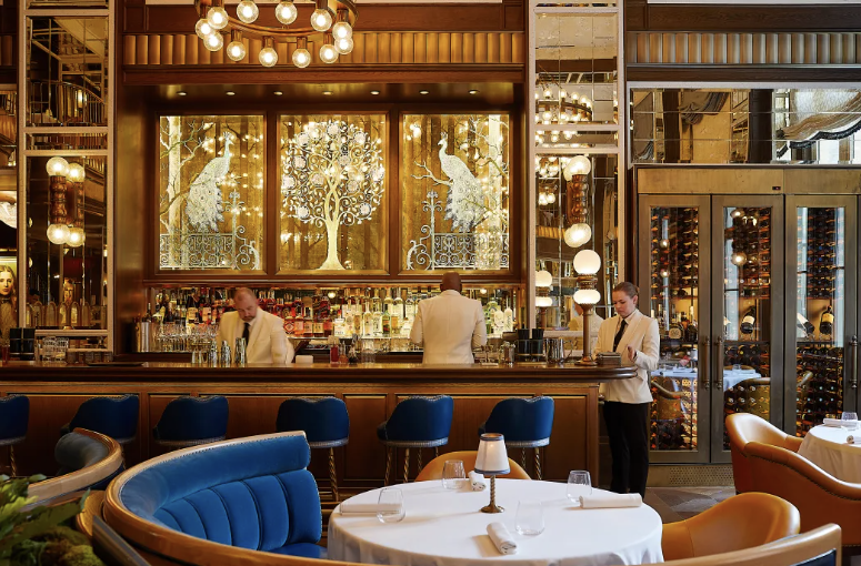 Elegant restaurant bar with blue and brown seating, three bartenders in white blazers, decorative wall art of peacocks and a tree with lights, warm lighting, and glass wall showcasing wine storage.