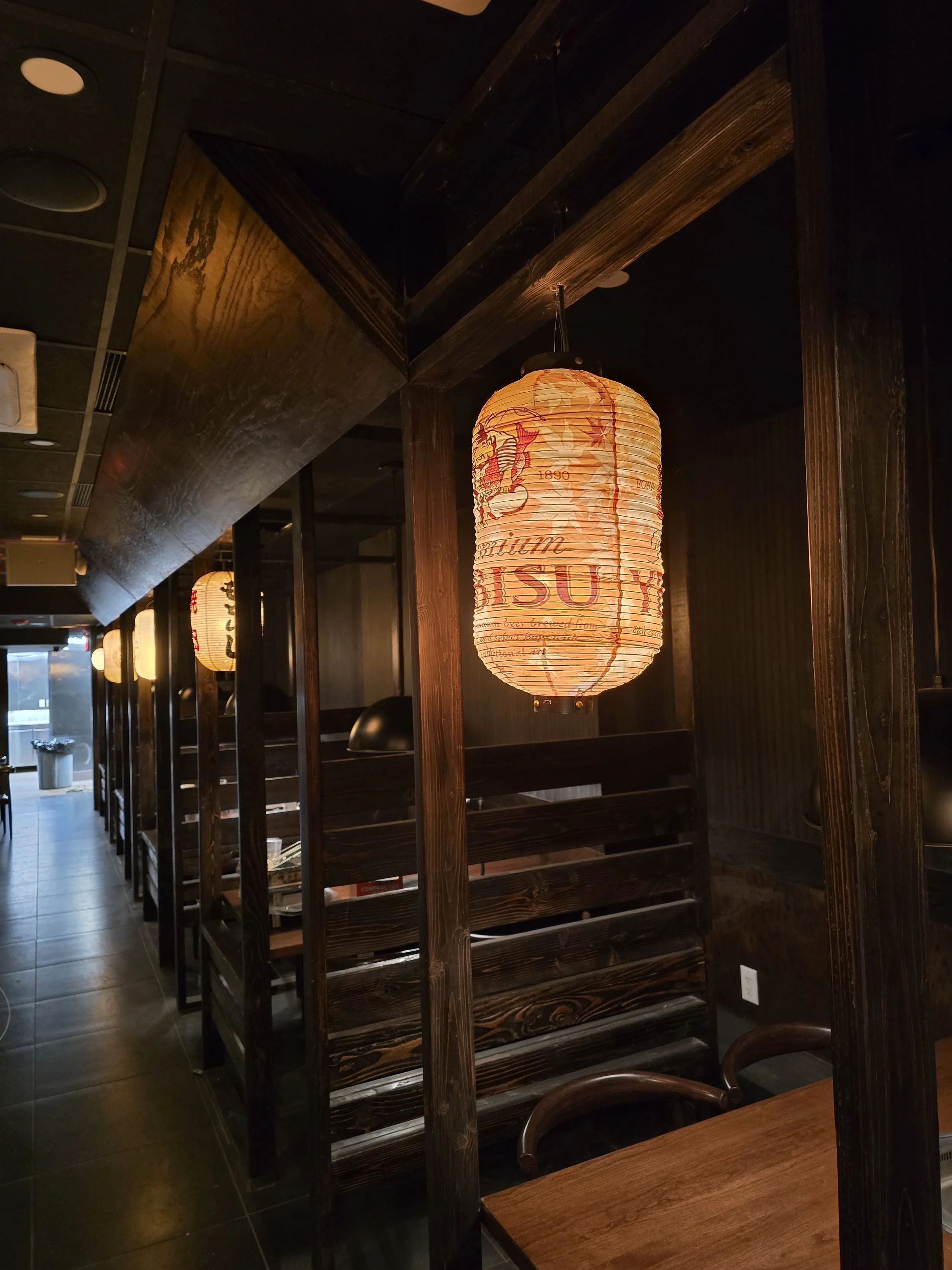 Dimly lit restaurant interior with wooden decor, hanging paper lanterns, and chairs.