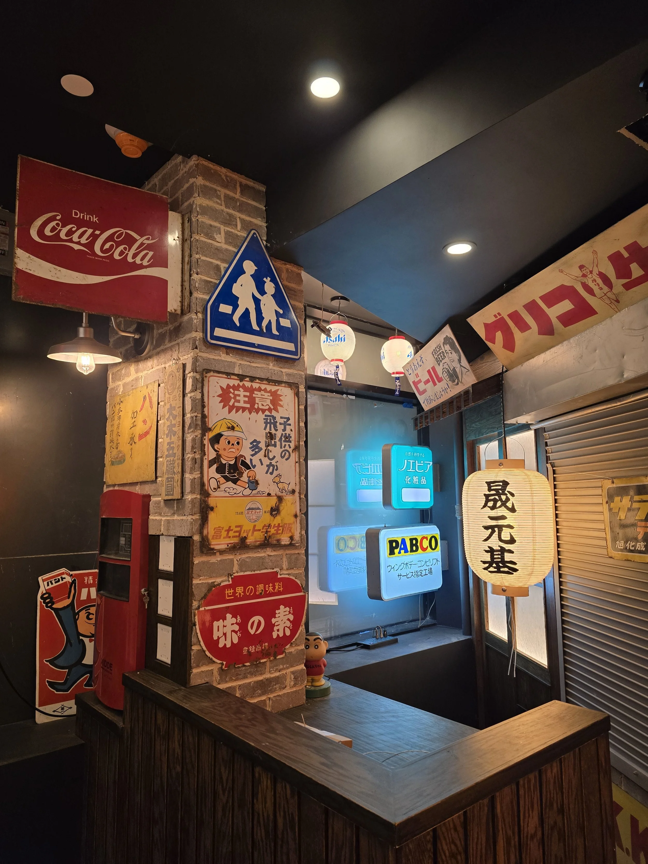 Interior of a Japanese-style restaurant or bar with vintage signs, lanterns, and neon lights, featuring a brick column and a dark ceiling.