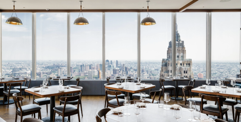 Upscale restaurant with floor-to-ceiling windows offering a city view, neatly set tables with glasses and cutlery, and modern pendant lighting.