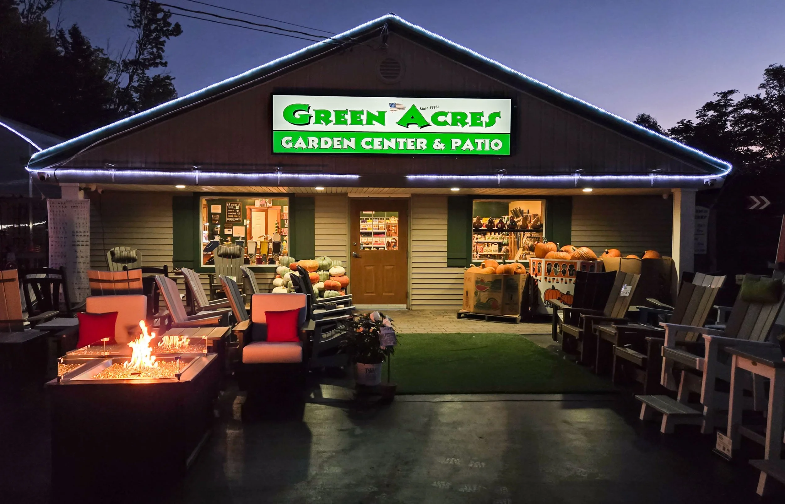 Green Acres Garden Center & Patio illuminated at dusk, with fire tables, outdoor seating and pumpkins displayed outside.
