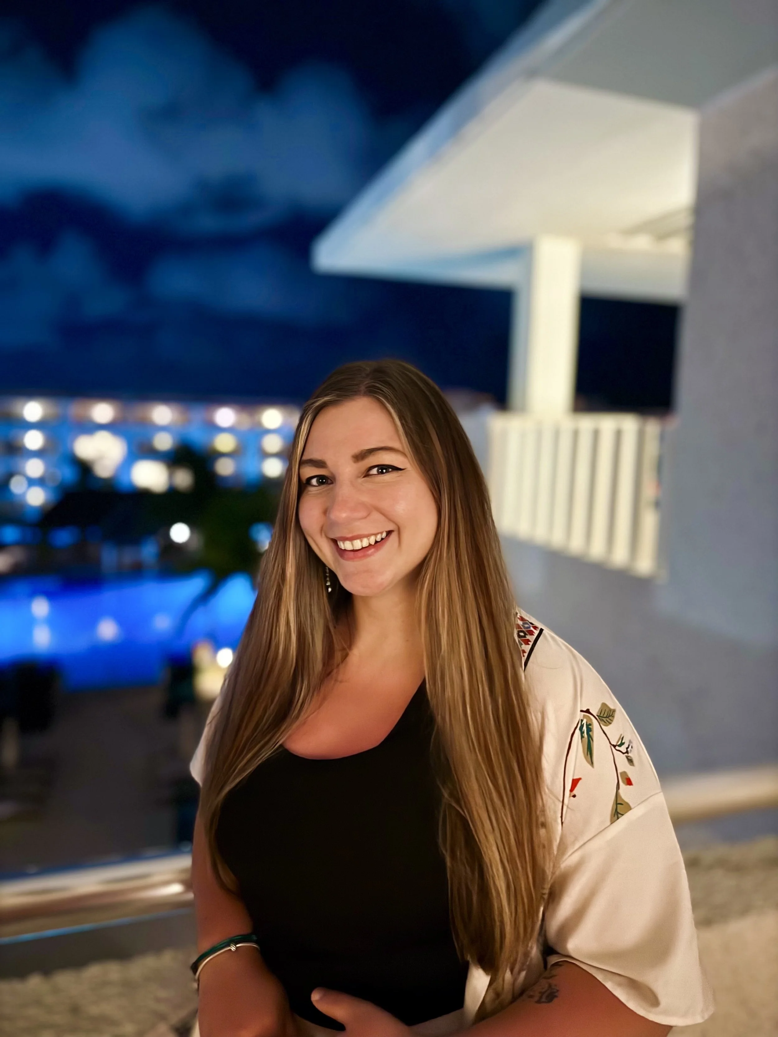 A woman with long brown hair smiling at night on a balcony with a lit pool and city buildings in the background.