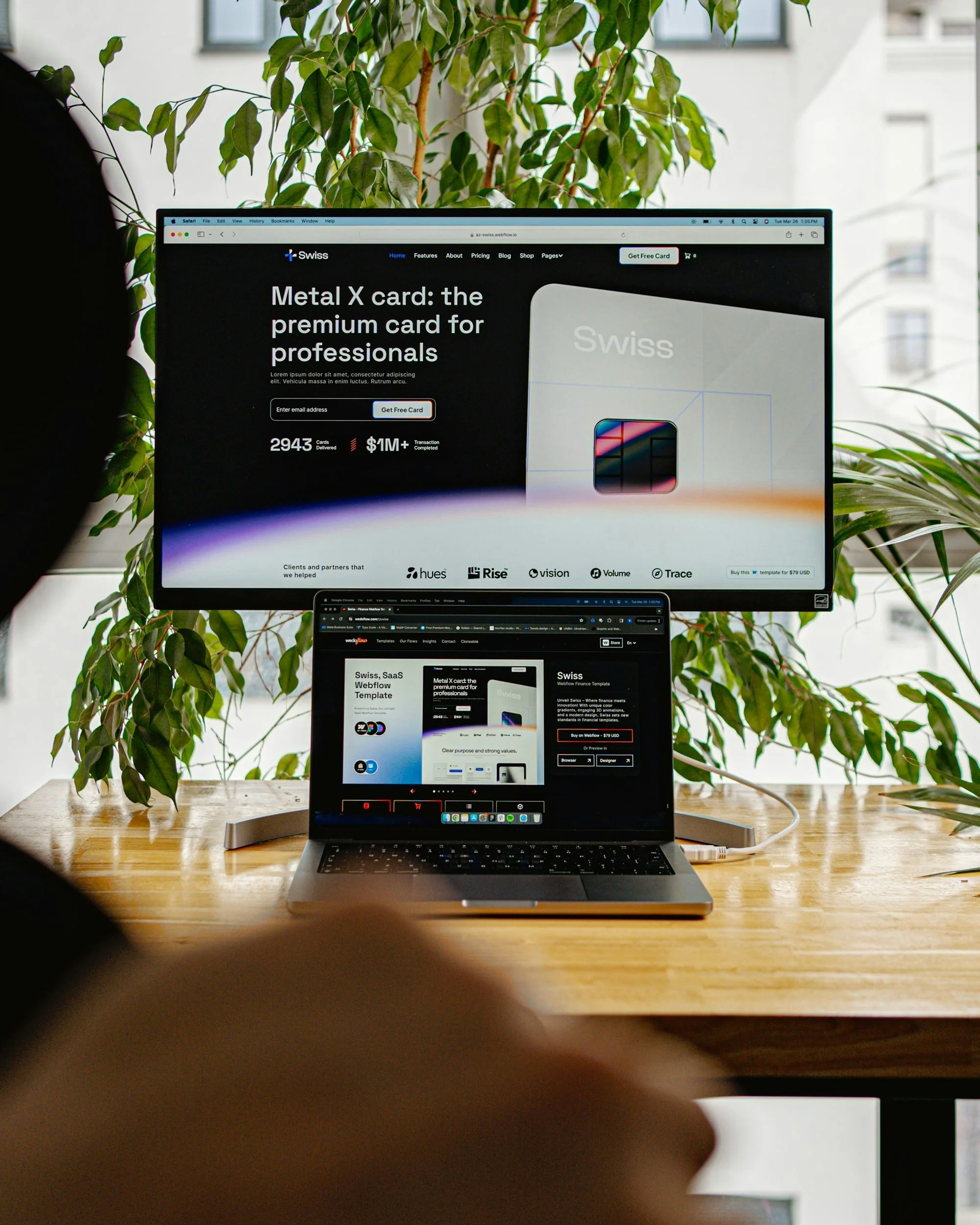 A workspace with a laptop on a wooden table displaying a website, and a large monitor above showing a webpage for Swiss Metal X card, with a background of green potted plants and windows.
