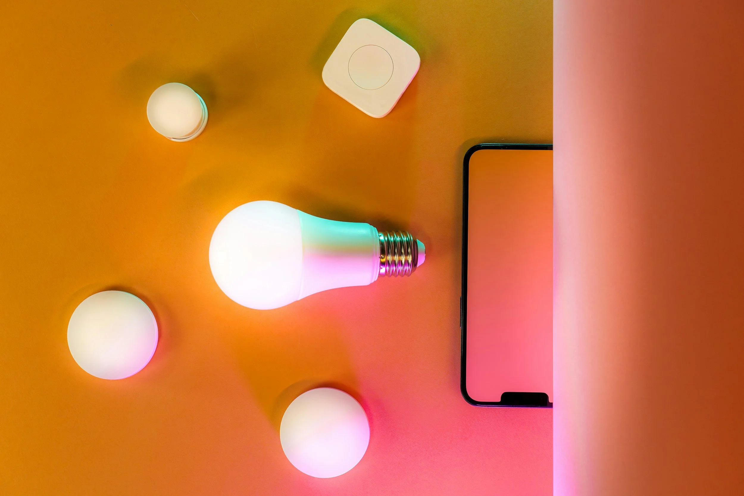 A digital photograph of a smartphone, a multicolored smart bulb, a white smart speaker, and a small white device on an orange and pink background.