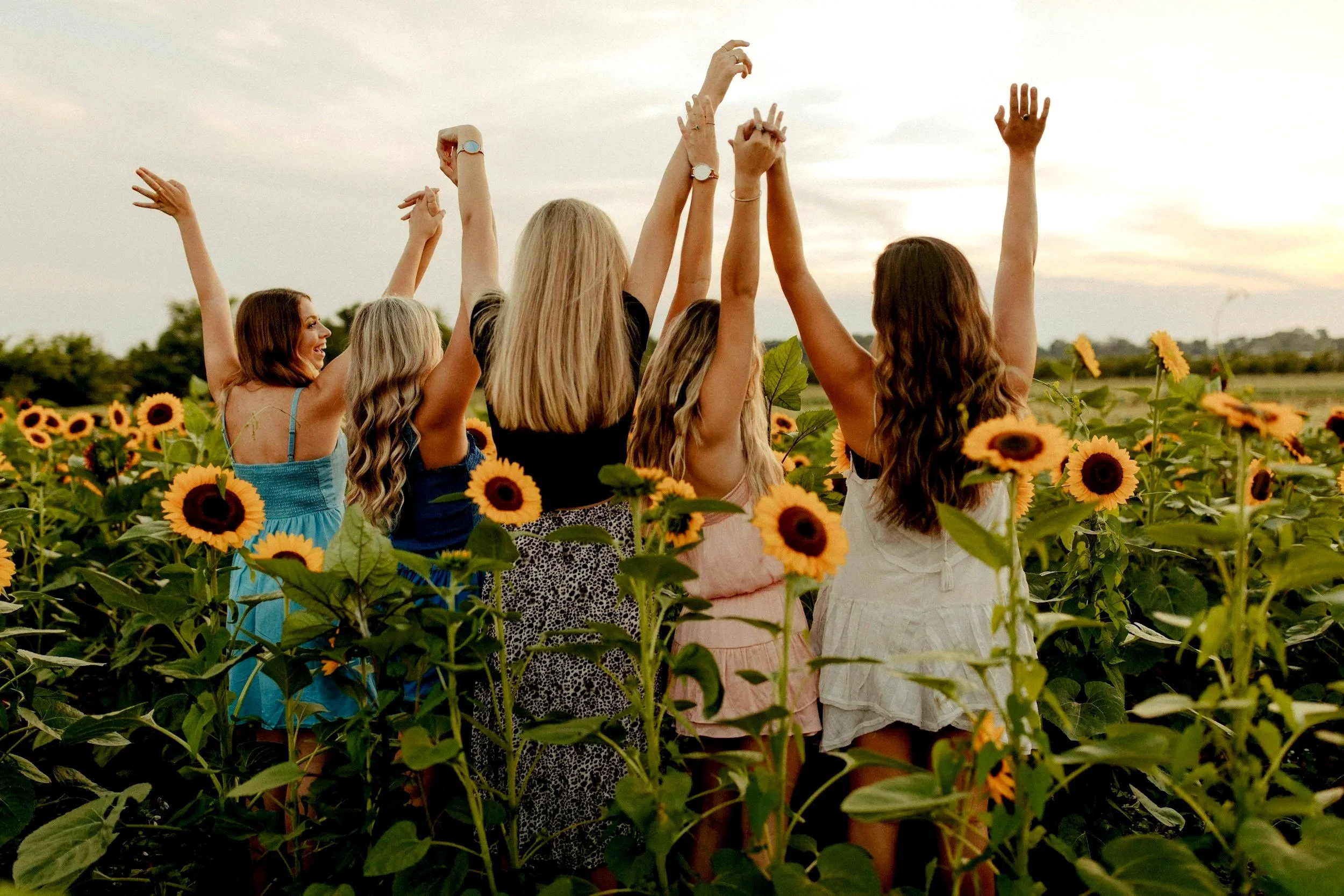 Five women standing in a sunflower field with their hands raised toward the sky during sunset, celebrating.