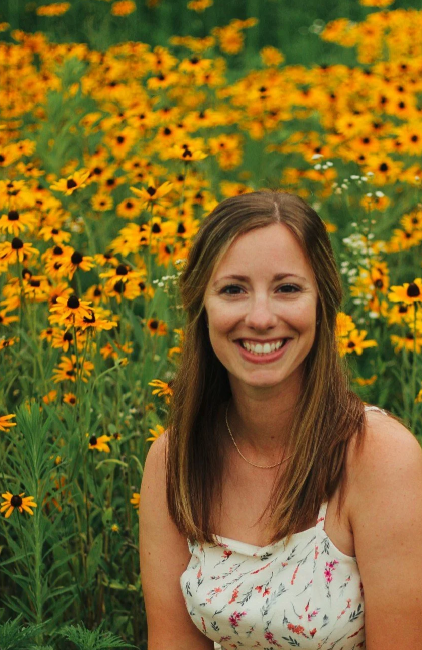 A woman with long brown hair smiling in a field of yellow and black flowers.
