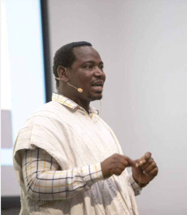 A man wearing a headset microphone delivers a presentation while gesturing.