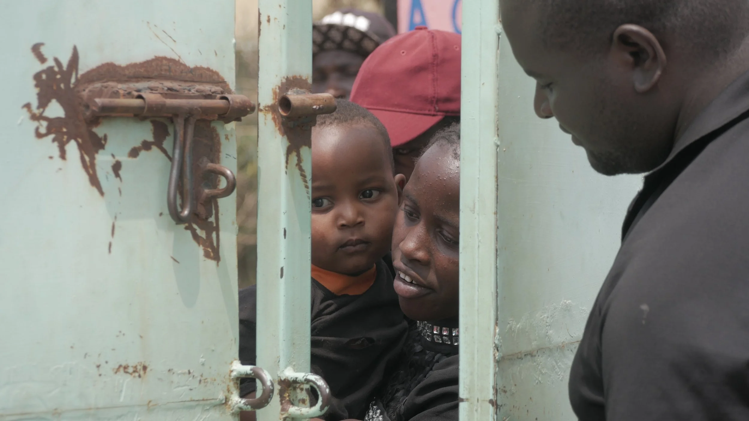 A caregiver holds a toddler beside a worn metal gate with an open padlock, while an adult man stands on the other side, reaching to open or close it.