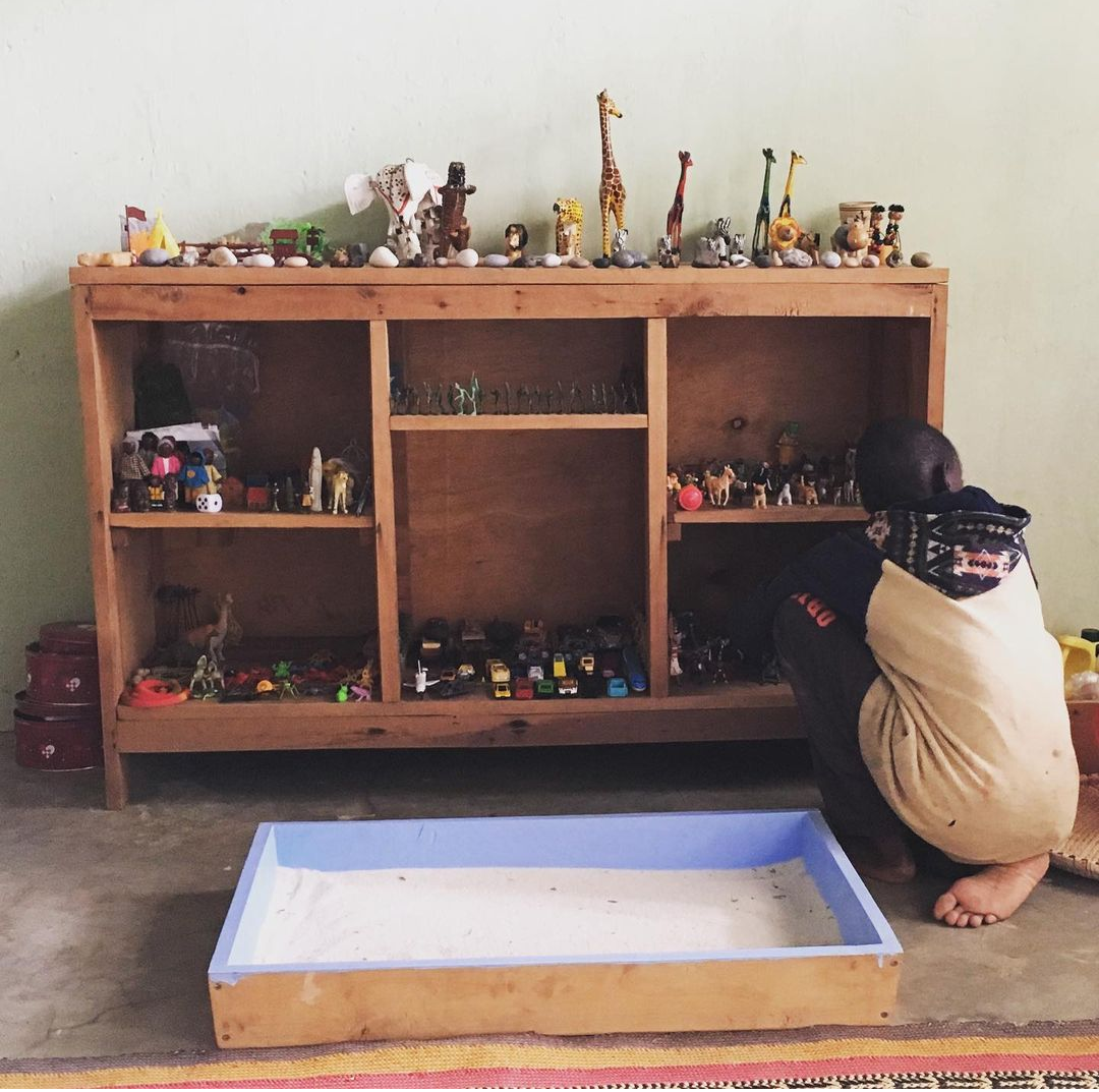 A child crouches beside a wooden shelf filled with small toy animals, figures, and vehicles with a tray filled with sand behind him.