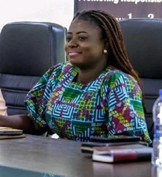 A woman with braided hair sits at a table in a meeting setting, smiling.