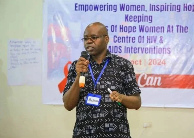 A man speaks into a microphone in front of a presentation backdrop with the words “Empowering Women” visible.
