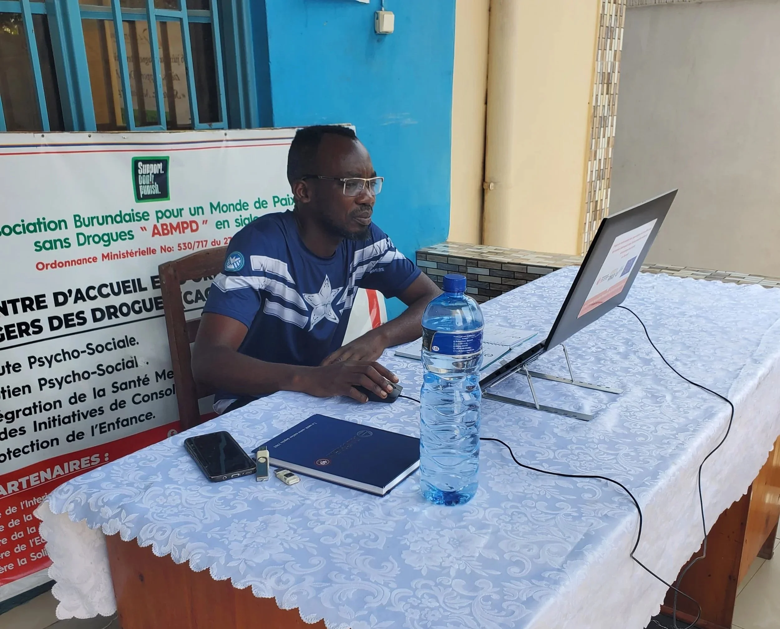 A man sits at a table outside a blue painted building working on a laptop, with a water bottle and notebook beside him.