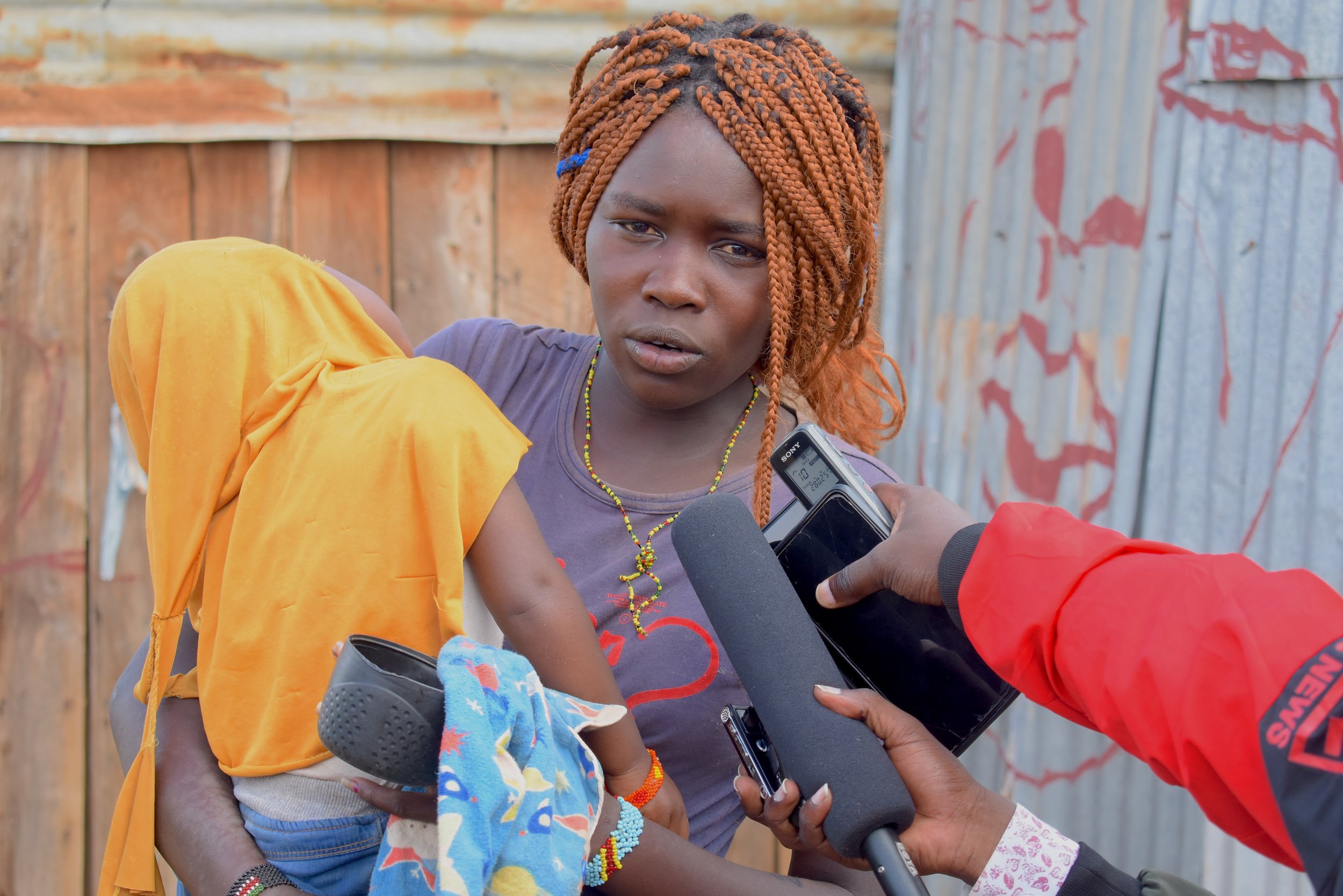 An adult woman holds a child on their hip while speaking, as multiple microphones and recording devices are held toward them in front of wooden and corrugated iron walls.