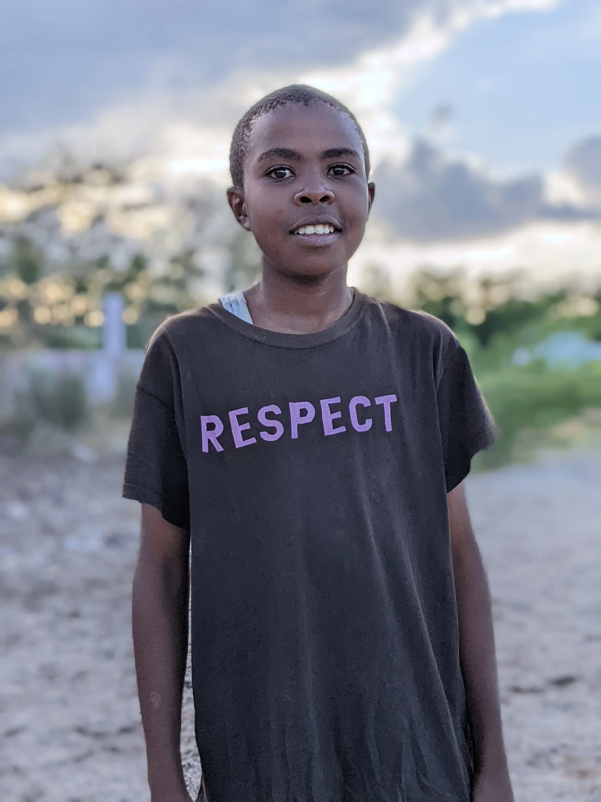 A child stands outdoors looking toward the camera, wearing a shirt with the word “Respect.”