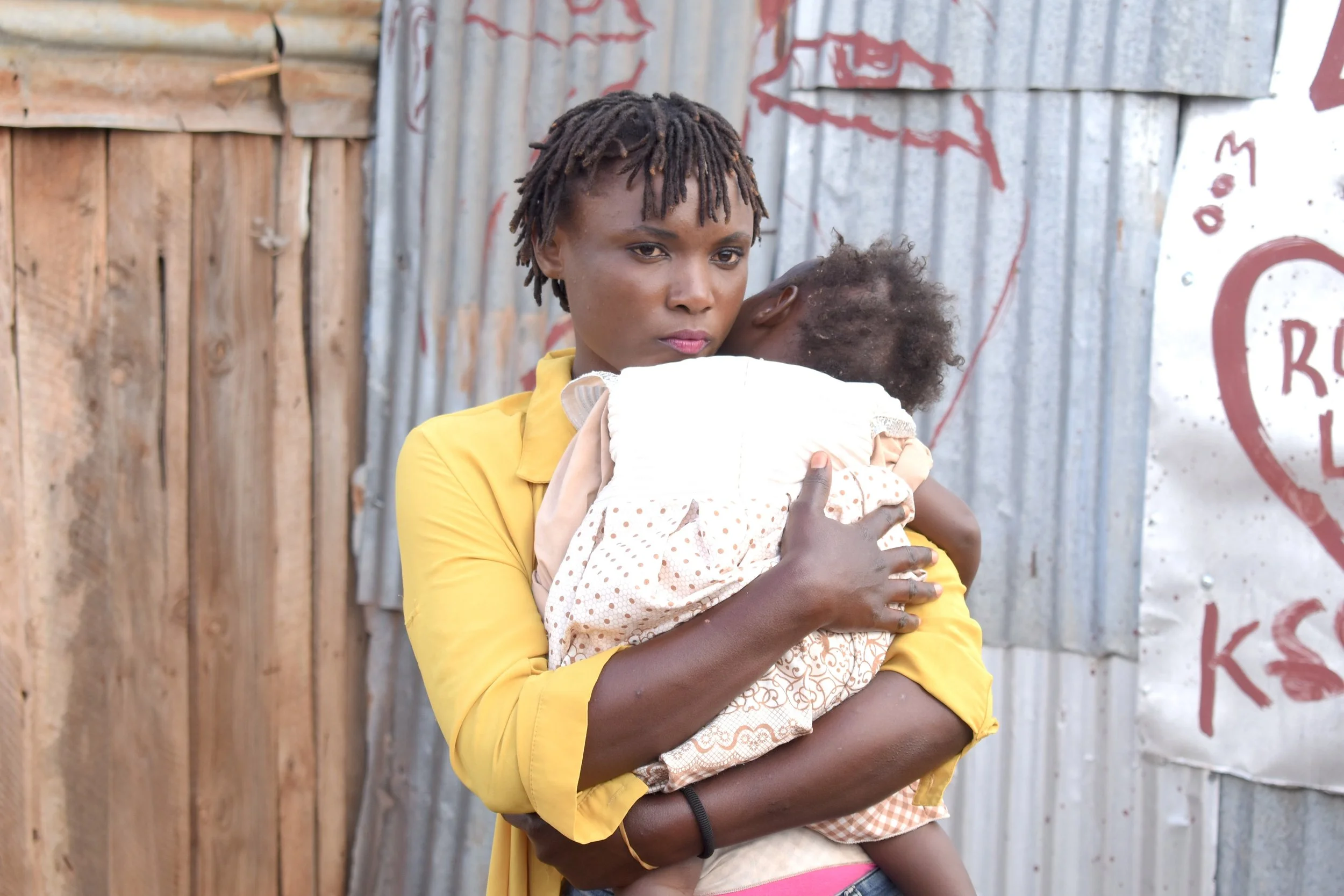 An adult woman cuddles a small child against their shoulder and looks preoccupied, standing in front of a corrugated metal building.