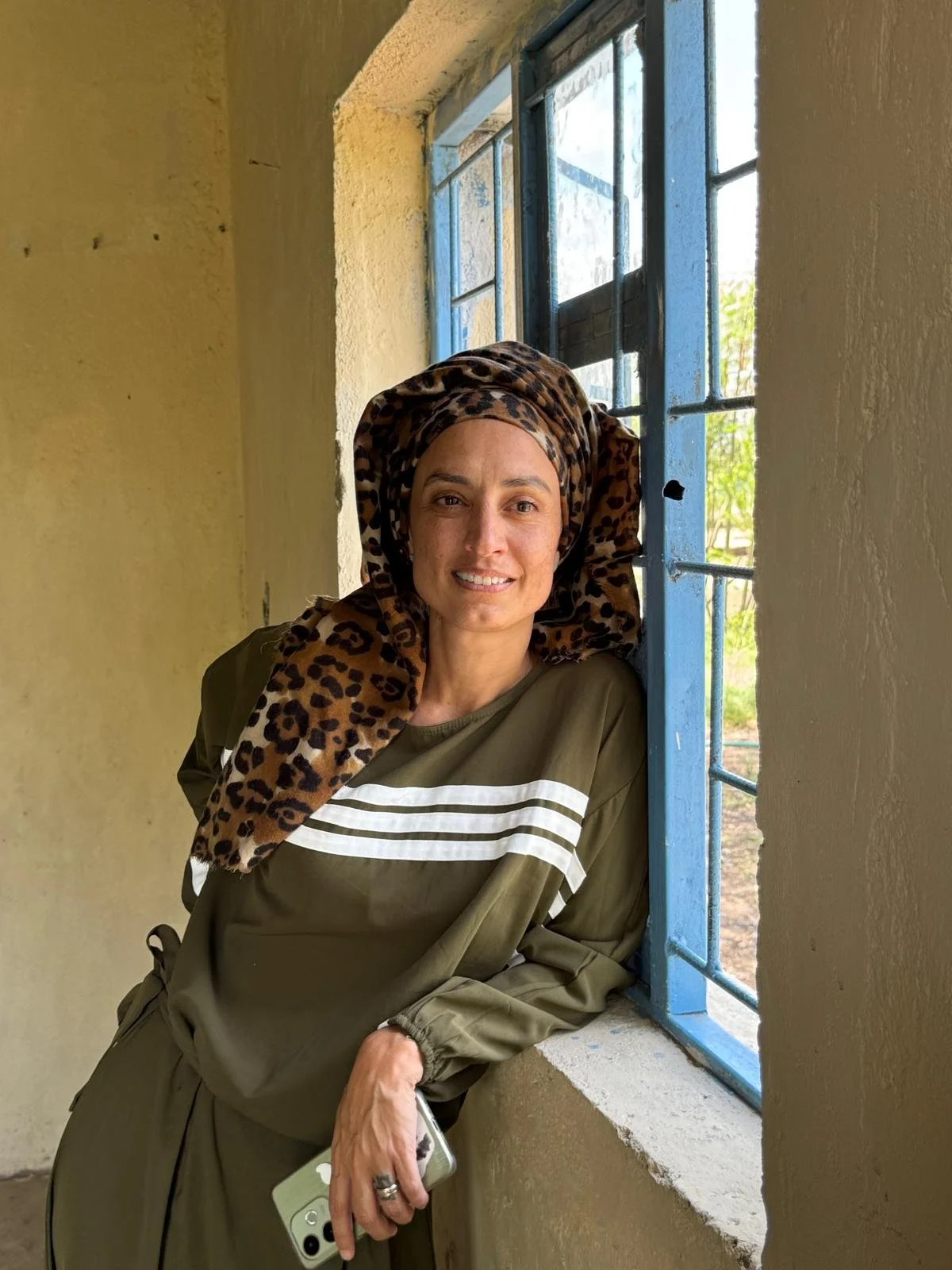 An adult woman wearing a patterned headscarf leans on a window ledge inside a yellow painted room, smiling toward the camera.